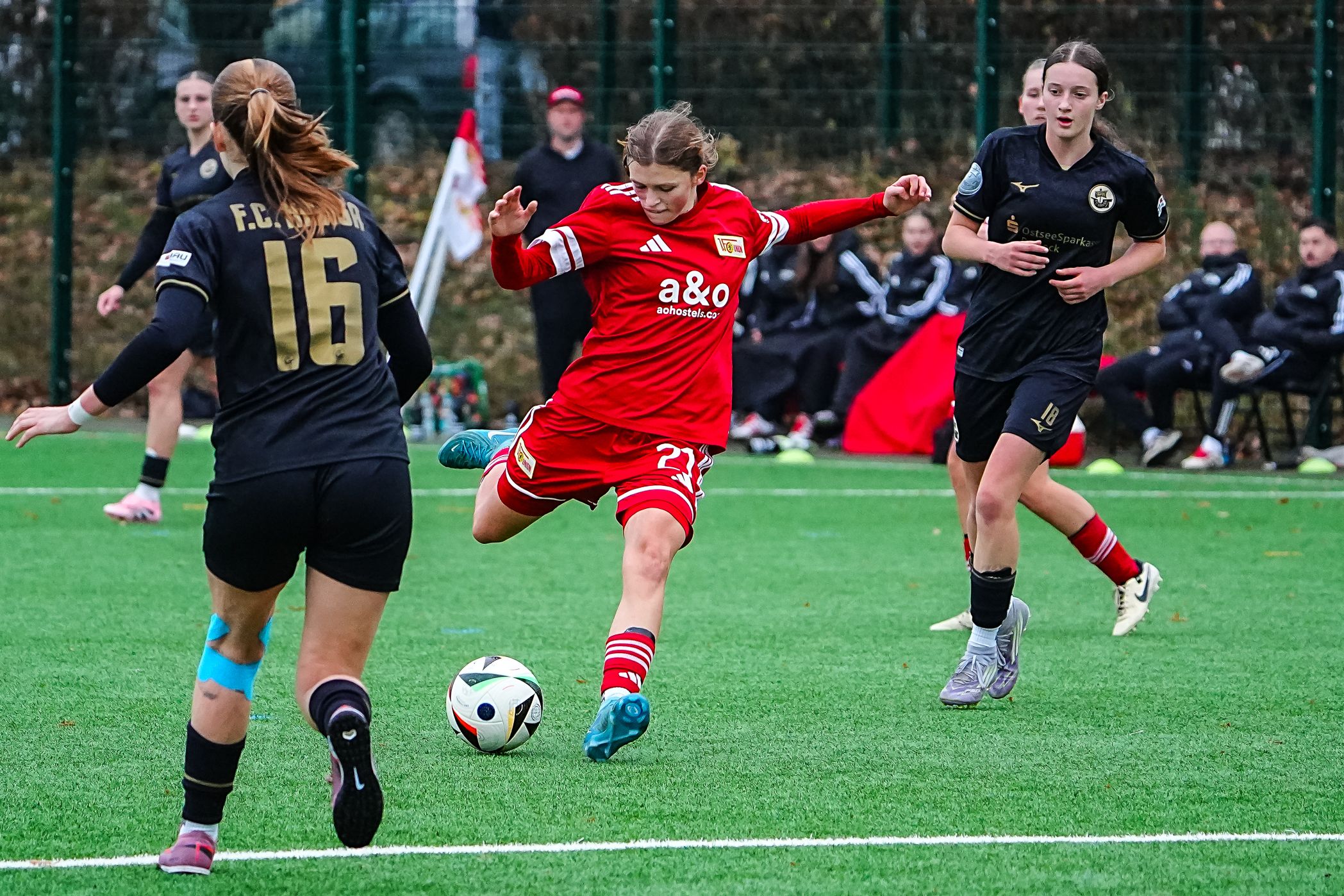 Eine Fußballspielerin in rotem Trikot schießt den Ball, während zwei Gegnerinnen ihm entgegenlaufen.