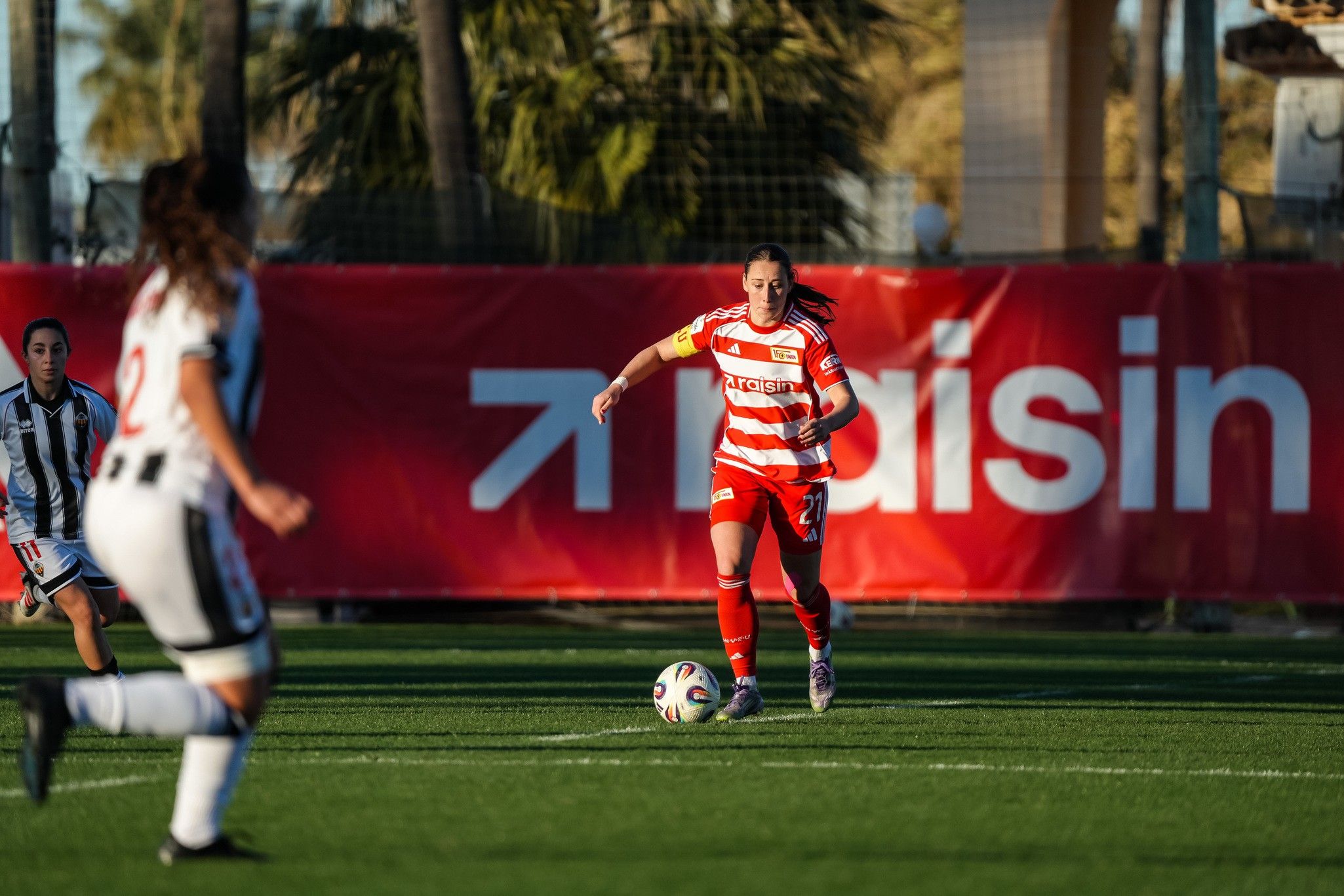 Eine Fußballspielerin in rotem Trikot dribbelt den Ball auf einem grünen Feld, während eine Gegnerin in Schwarz-Weiß im Hintergrund steht.