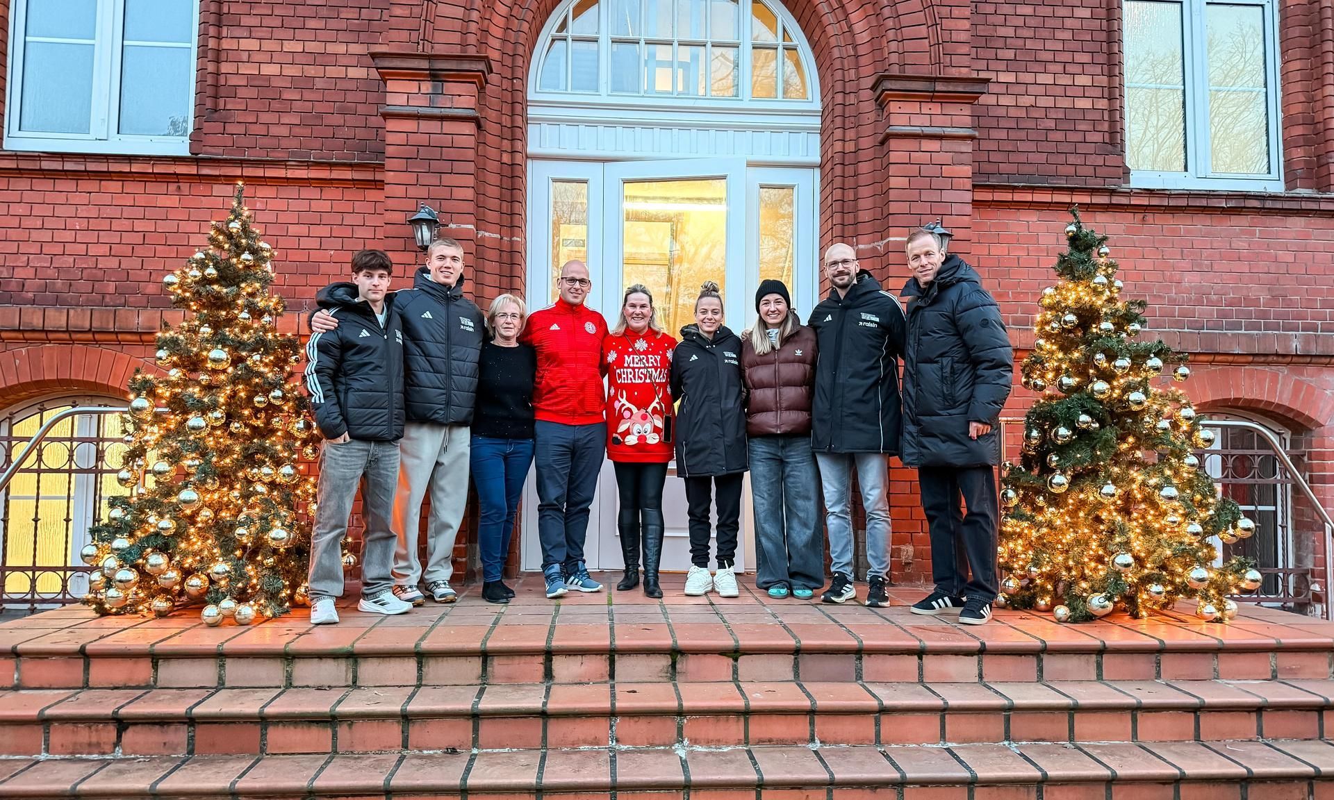 Eine Gruppe von neun Personen steht vor einem Gebäude, umgeben von festlich geschmückten Weihnachtsbäumen mit Lichtern.