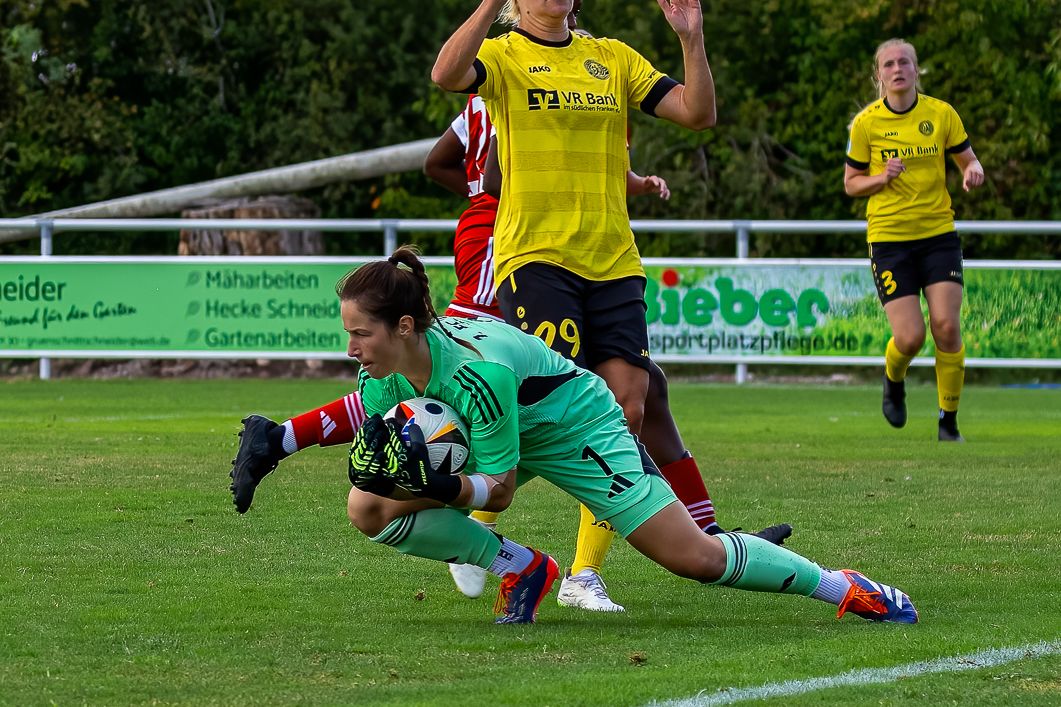 Eine Torhüterin in grüner Ausrüstung fängt den Ball, während eine Spielerin im gelben Trikot im Hintergrund steht.