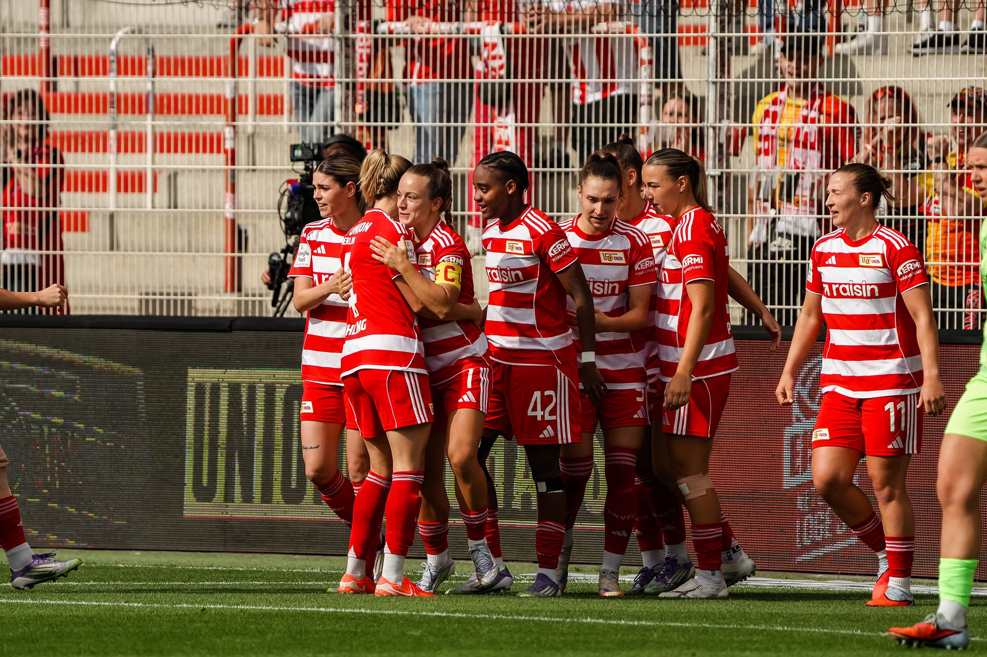 Eine Gruppe von Fußballspielerinnen in roten und weißen Trikots feiert einen Torerfolg auf dem Spielfeld.