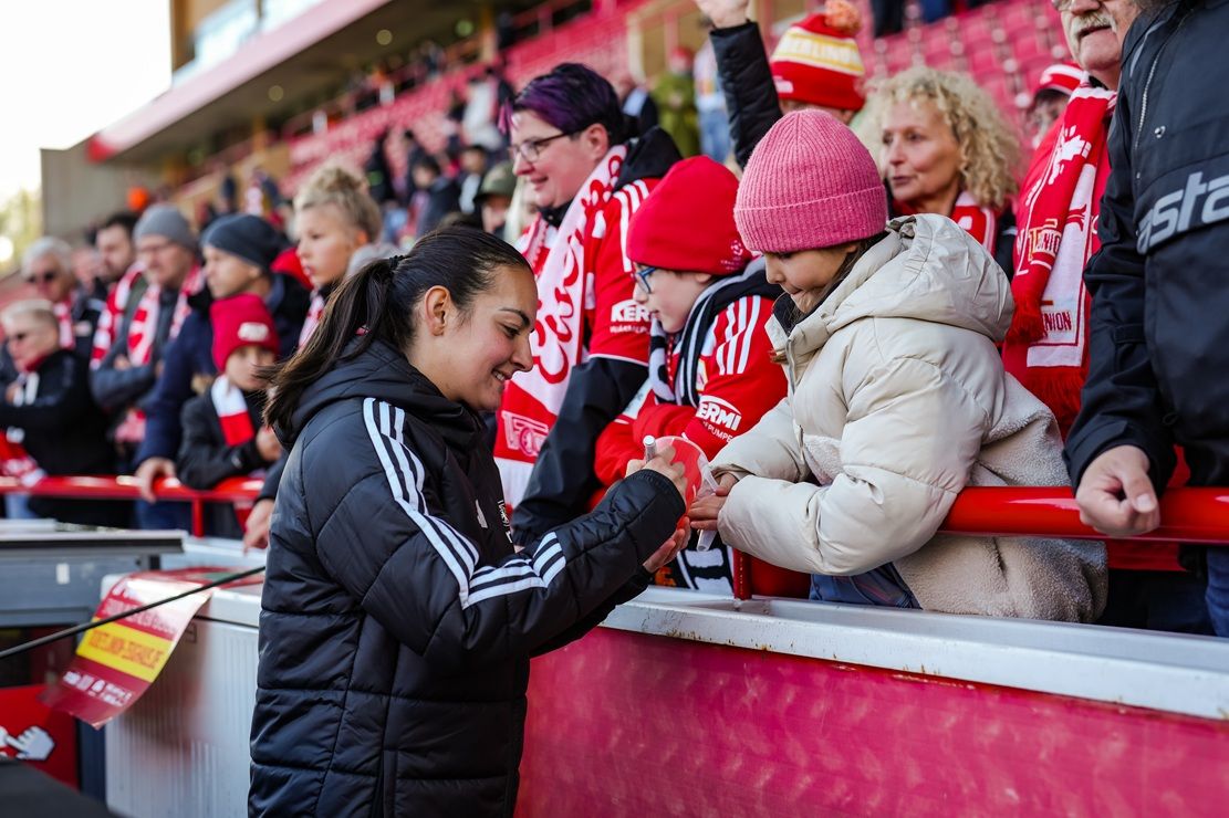 Eine Sportlerin interagiert lächelnd mit Fans, während diese in einem Stadion stehen, umgeben von Fanfare und Teamfarben.