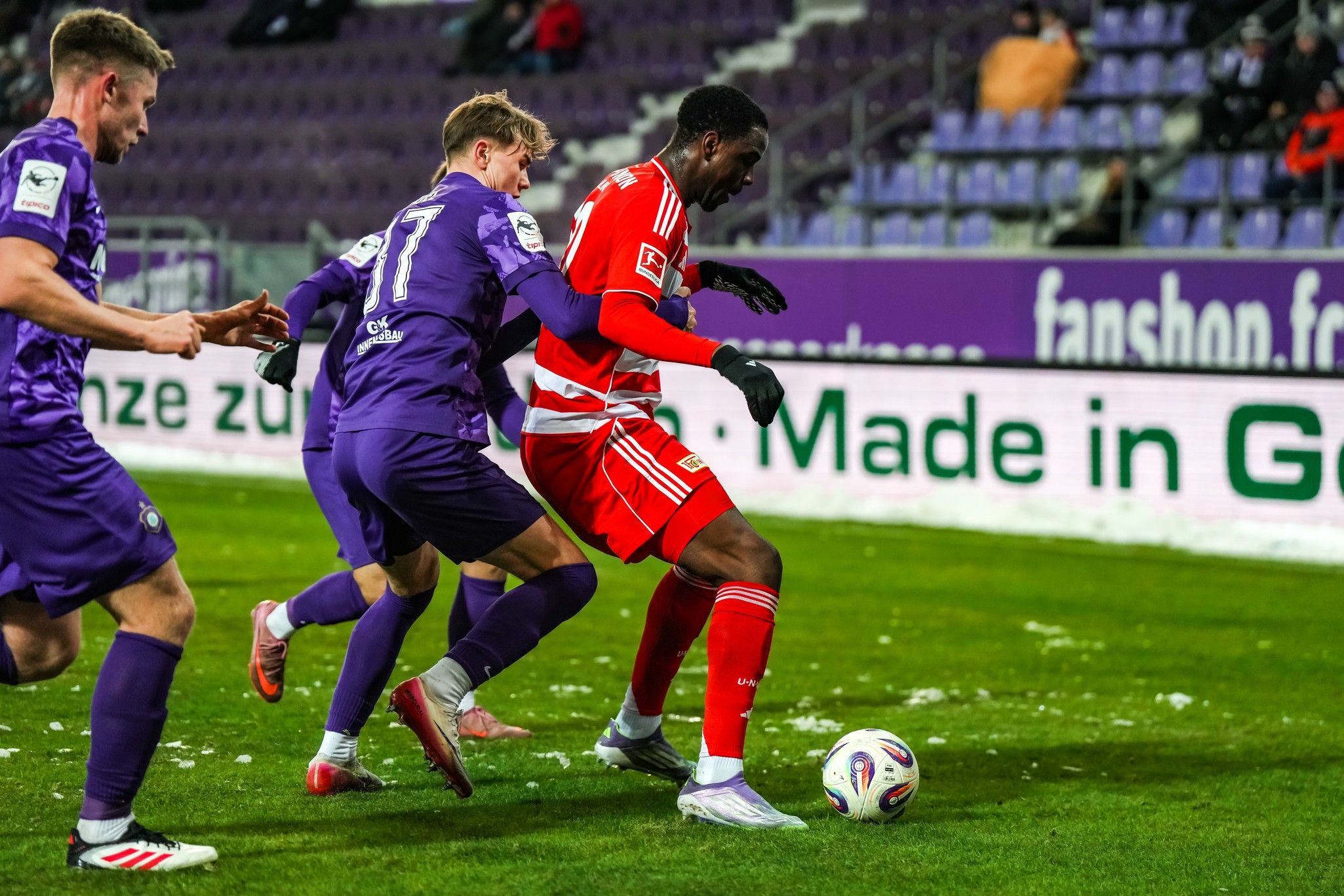 Zwei Fußballspieler in Lila versuchen, einen dritten Spieler in Rot am Ball zu hindern, während sie auf einem grünen Spielfeld stehen.