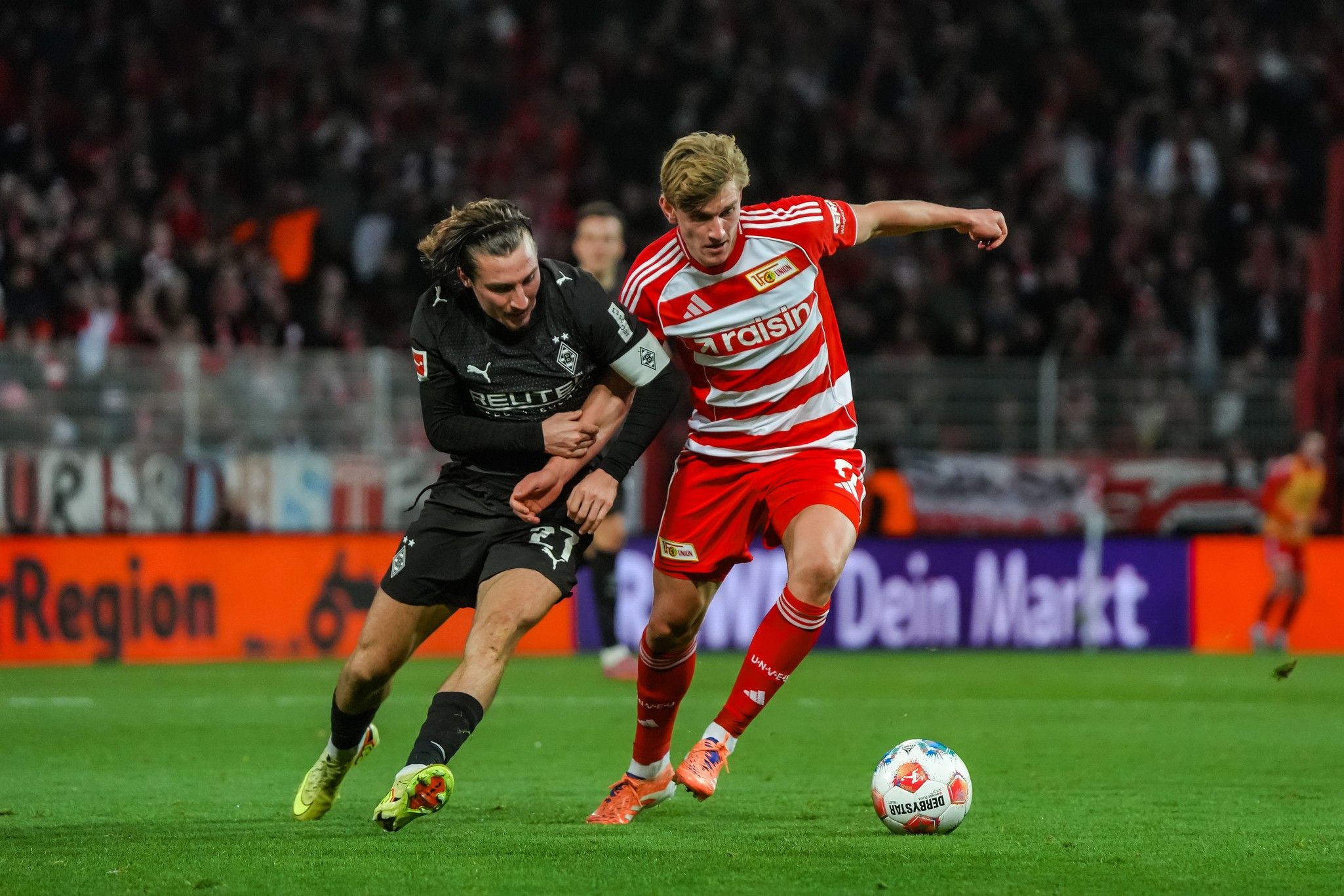 Fußballspiel zwischen zwei Spielern, einer in schwarzer und der andere in rot-weißer Uniform, auf einem Stadionrasen.