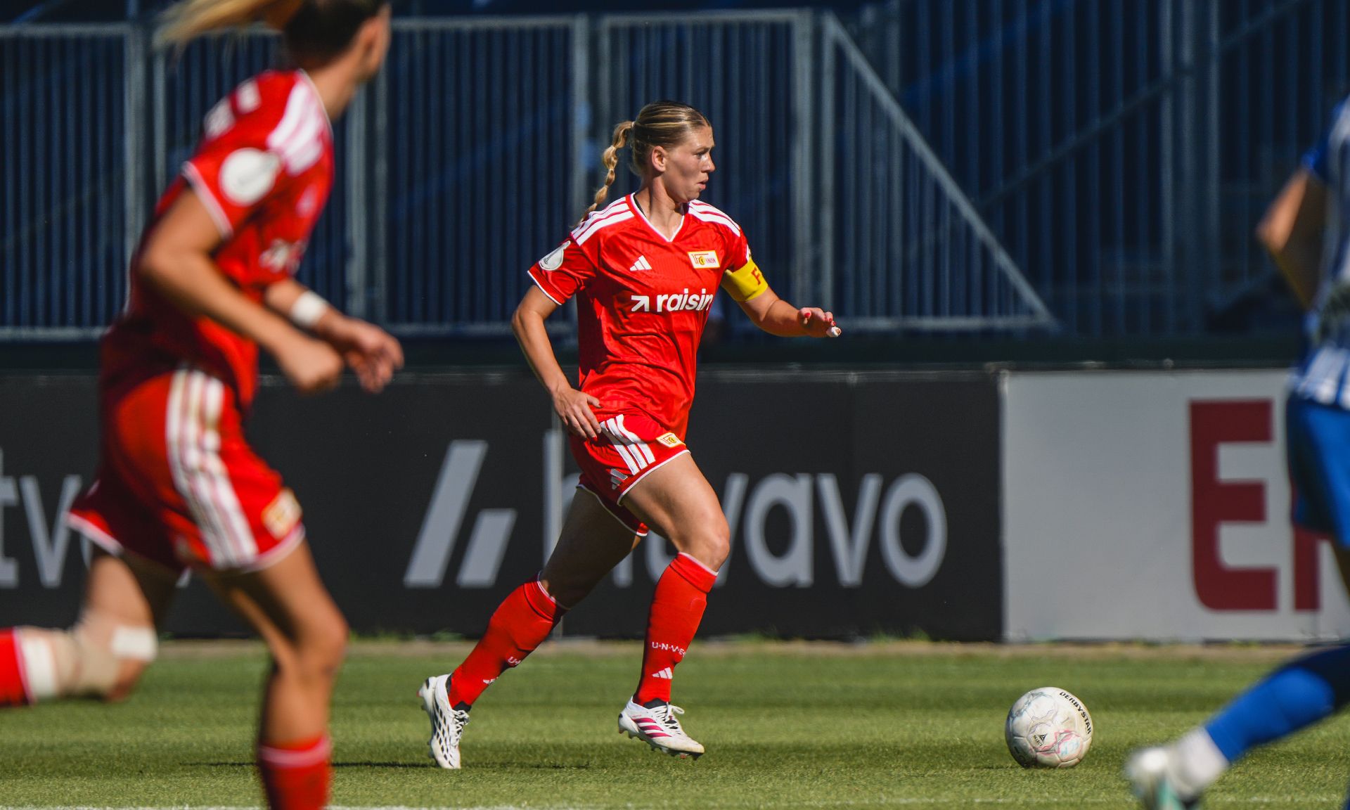 Eine Spielerin in rotem Trikot mit der Nummer 7 dribbelt den Ball auf dem Spielfeld, während andere Spielerinnen im Hintergrund agieren.