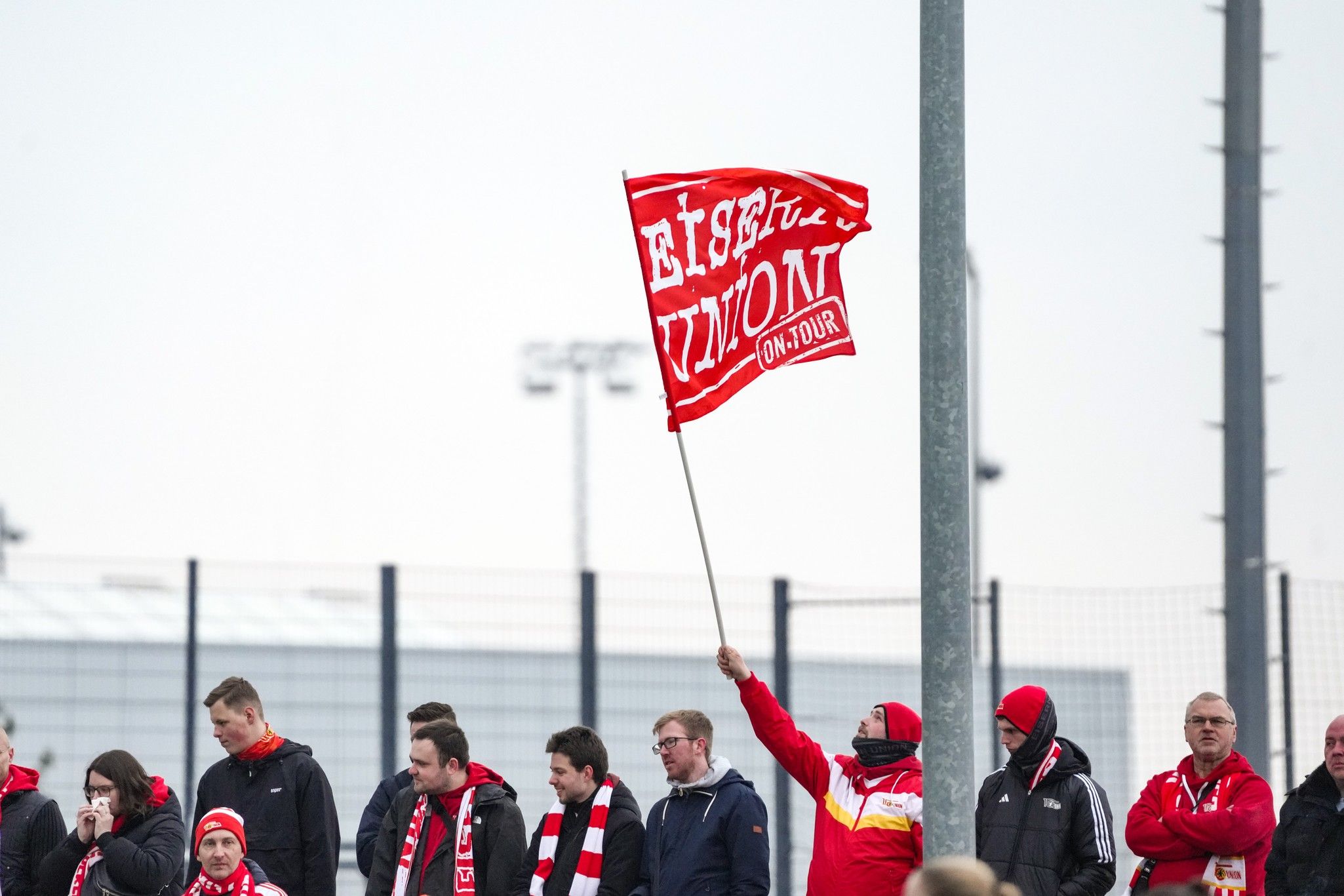 Ein Fan hält eine rote Flagge mit der Aufschrift „EISERN UNION“ während eines Fußballspiels, umgeben von weiteren Anhängern.
