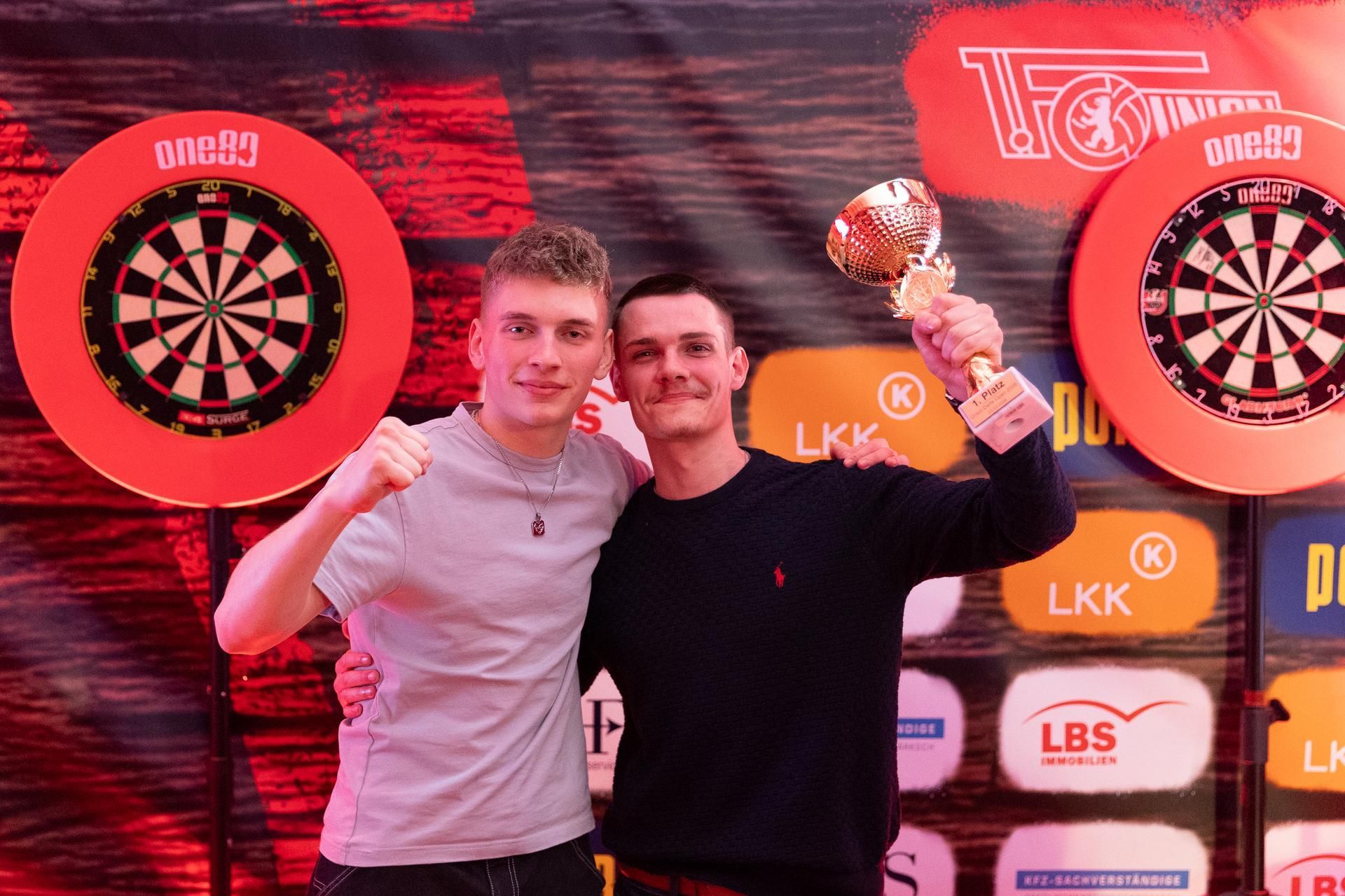 Zwei junge Männer posieren lächelnd für die Kamera, einer hält einen Pokal, im Hintergrund Dartscheiben und bunte Lichtdekoration.