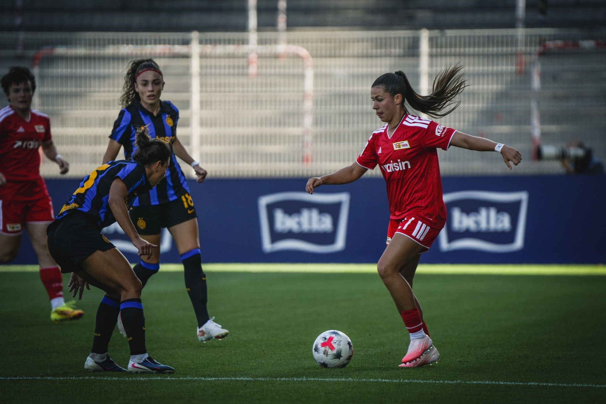 Eine Fußballspielerin in roter Ausrüstung dribbelt den Ball, während zwei gegnerische Spielerinnen in blauen Trikots nahe sind.