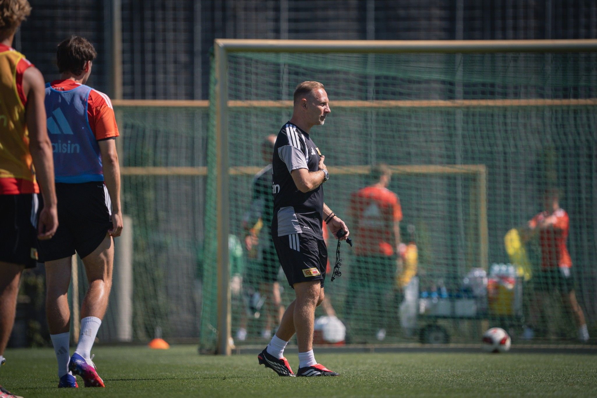 Ein Trainer beobachtet Spieler während eines Fußballtrainings am Platz mit Toren im Hintergrund.
