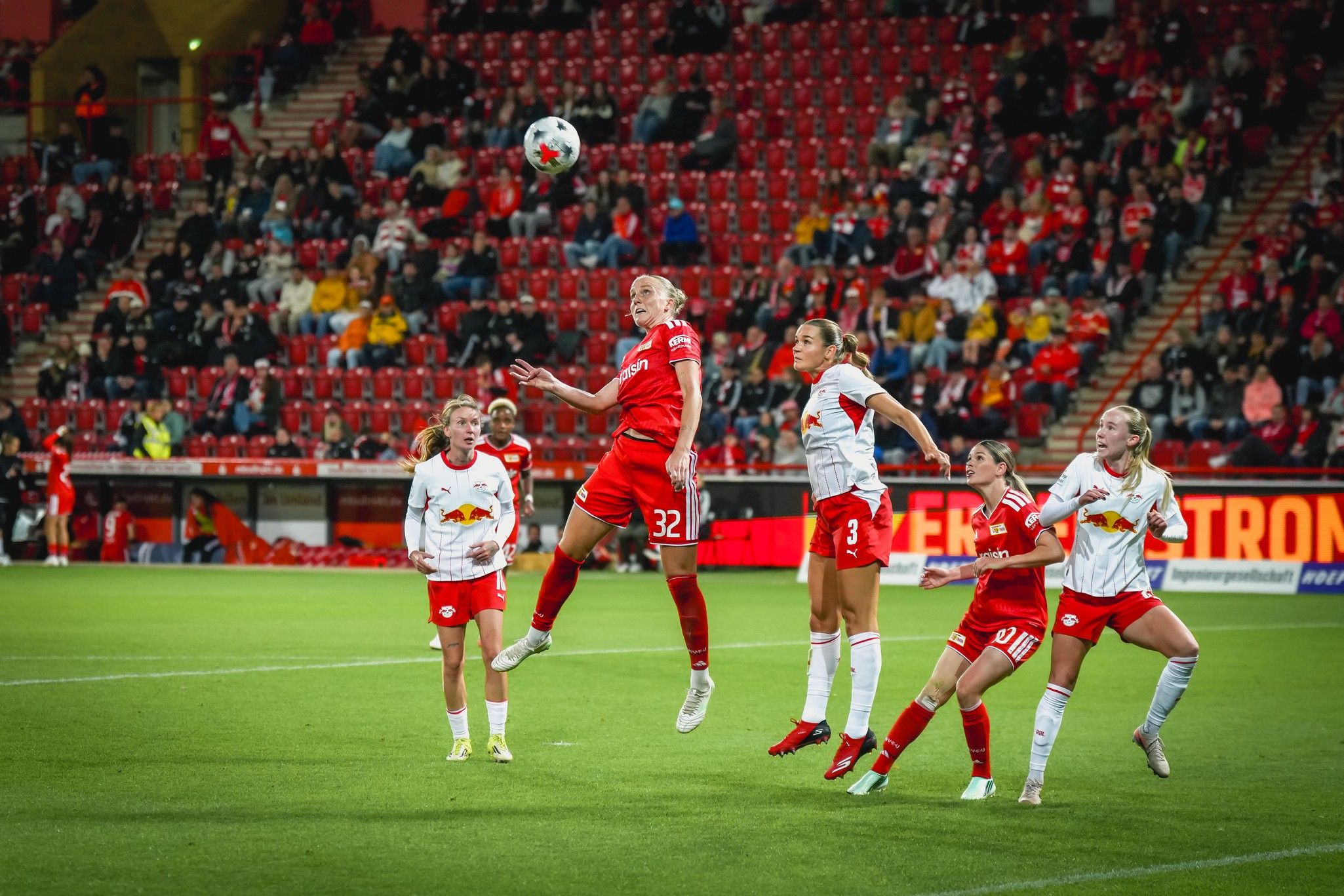 Spielszene aus einem Fußballspiel: Eine Spielerin springt hoch, während andere Spielerinnen springen oder sich positionieren. Zuschauer im Hintergrund.
