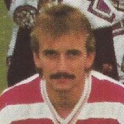 Ein Mann mit einem Schnurrbart und einem gestreiften Trikot schaut direkt in die Kamera. Raisinträgers hinter ihm.