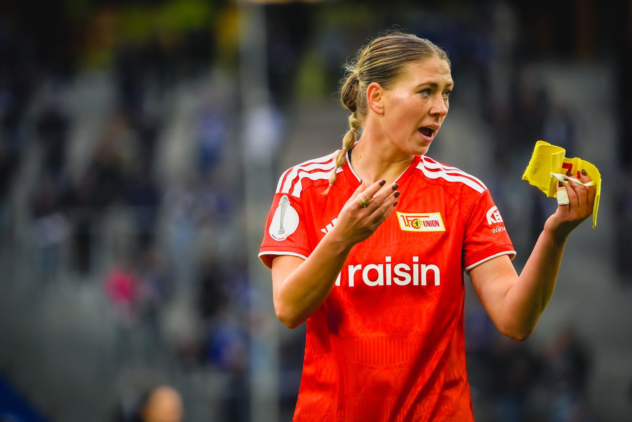 Fußballspielerin in rotem Trikot hält gelbe Karten in der Hand und kommuniziert auf dem Spielfeld.