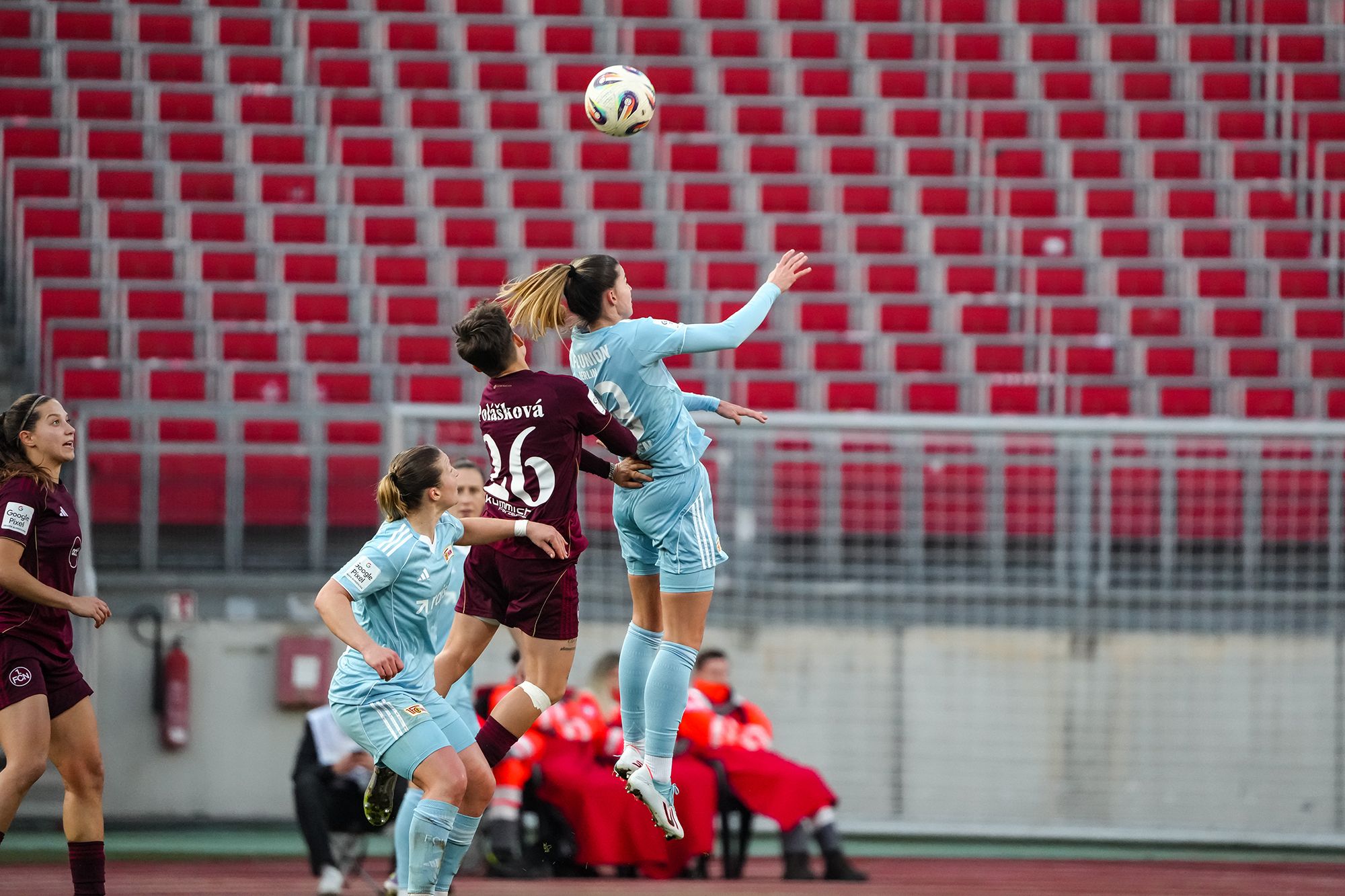 Zwei Fußballspielerinnen springen hoch, um den Ball zu erreichen, während Zuschauer im Hintergrund zu sehen sind.