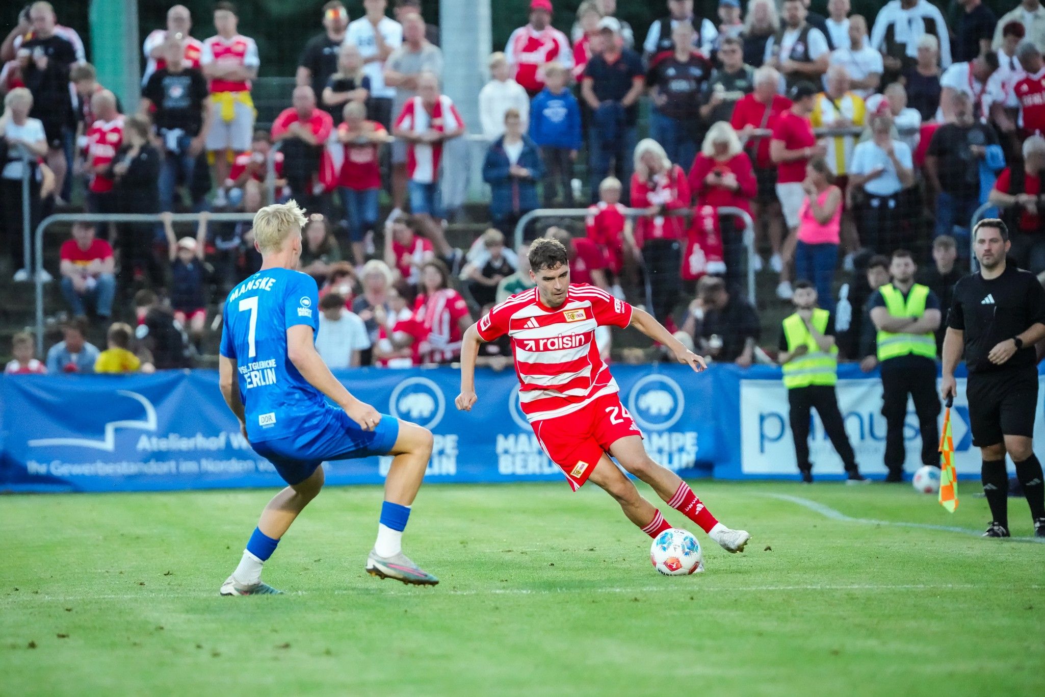 Fußballspiel zwischen zwei Mannschaften auf einem Rasenfeld, mit Spielern in roten und blauen Trikots und Zuschauern im Hintergrund.
