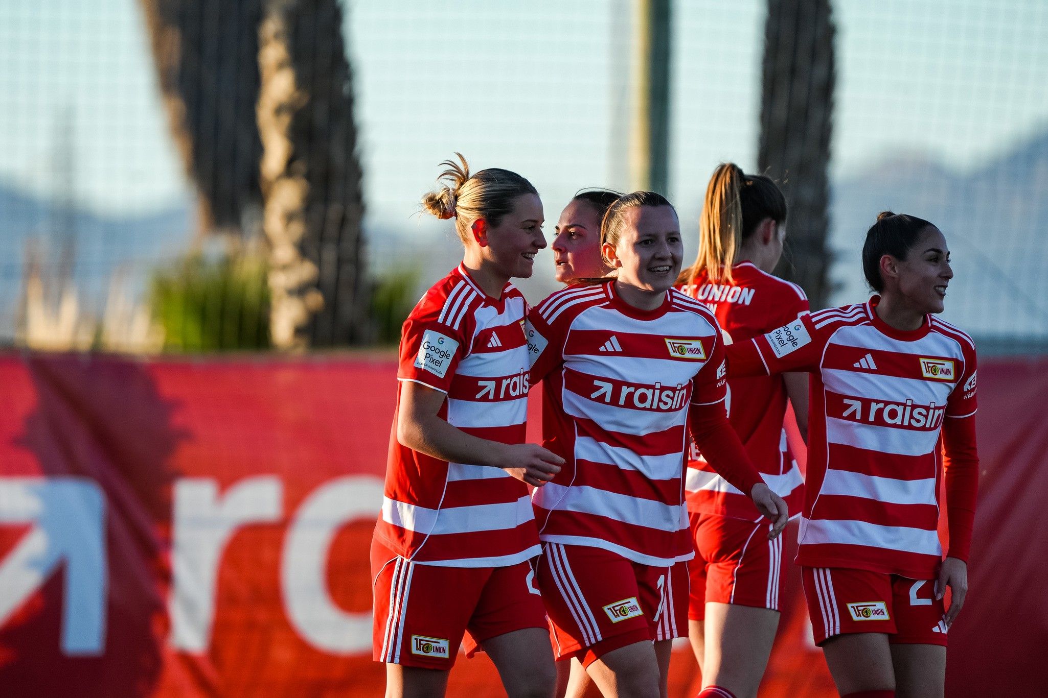 Zwei Fußballspielerinnen in roten Trikots lachen und feiern gemeinsam auf dem Spielfeld, während andere Spielerinnen im Hintergrund stehen.