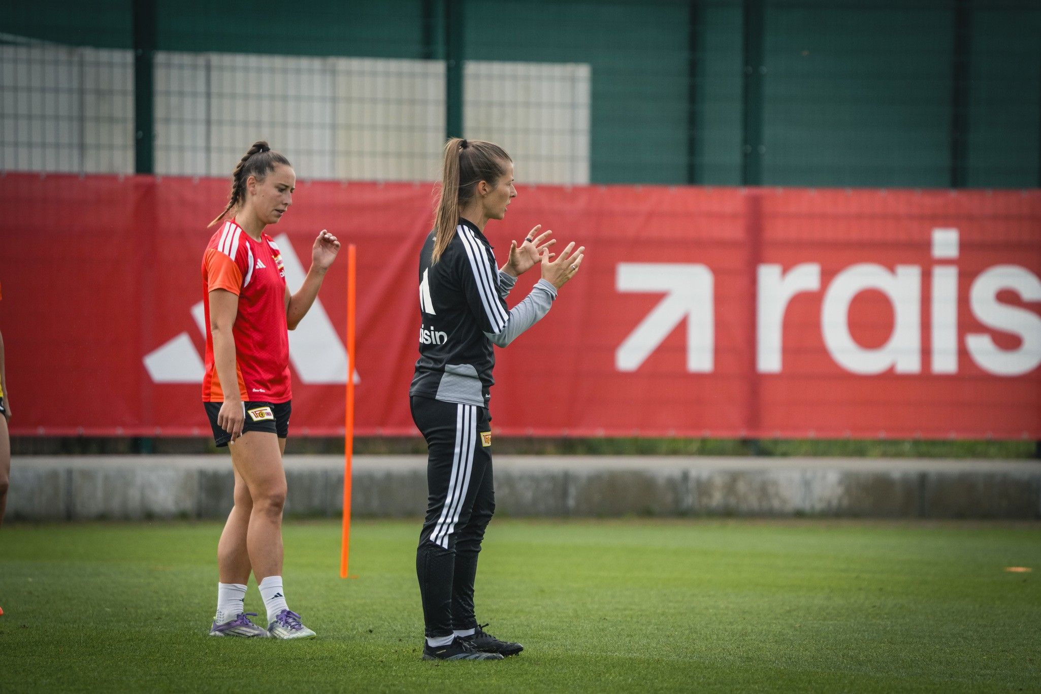 Zwei Fußballspielerinnen trainieren auf einem Platz, eine in rotem Trikot und eine in grauem Trainingsanzug, mit Markierungen und Platz im Hintergrund.