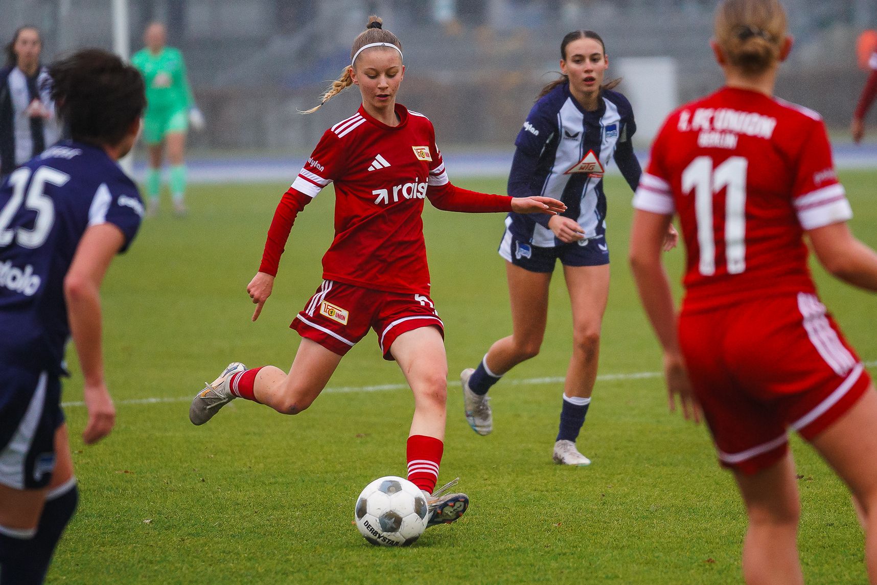 Eine Spielerin in roten Sporttrikots dribbelt den Fußball auf einem Kunstrasenplatz, während andere Spielerinnen in blauen und weißen Trikots beobachten.