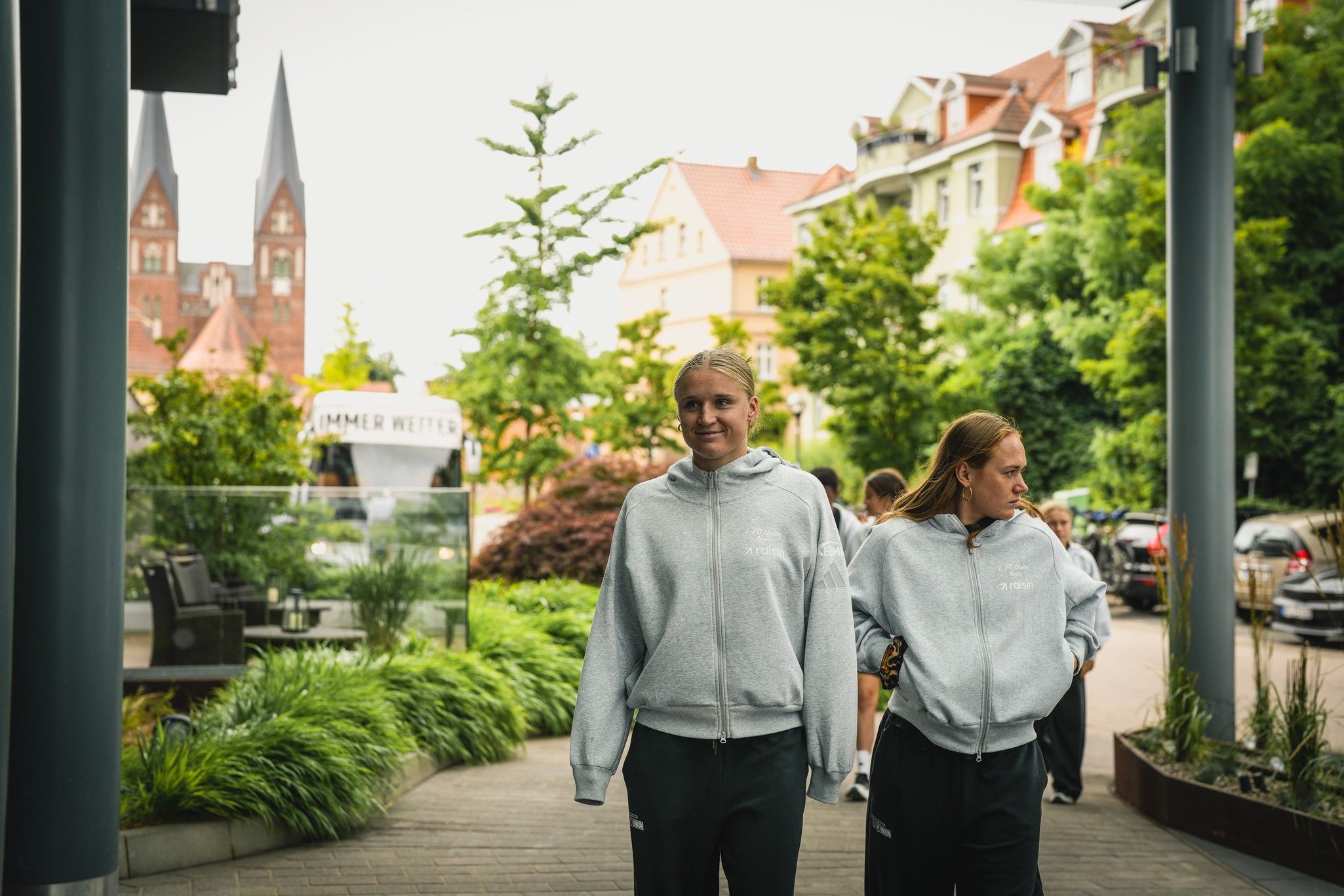 Zwei Personen in grauen Sweatshirts gehen an einer Straße entlang, umgeben von Bäumen und Gebäuden mit Türmen im Hintergrund.
