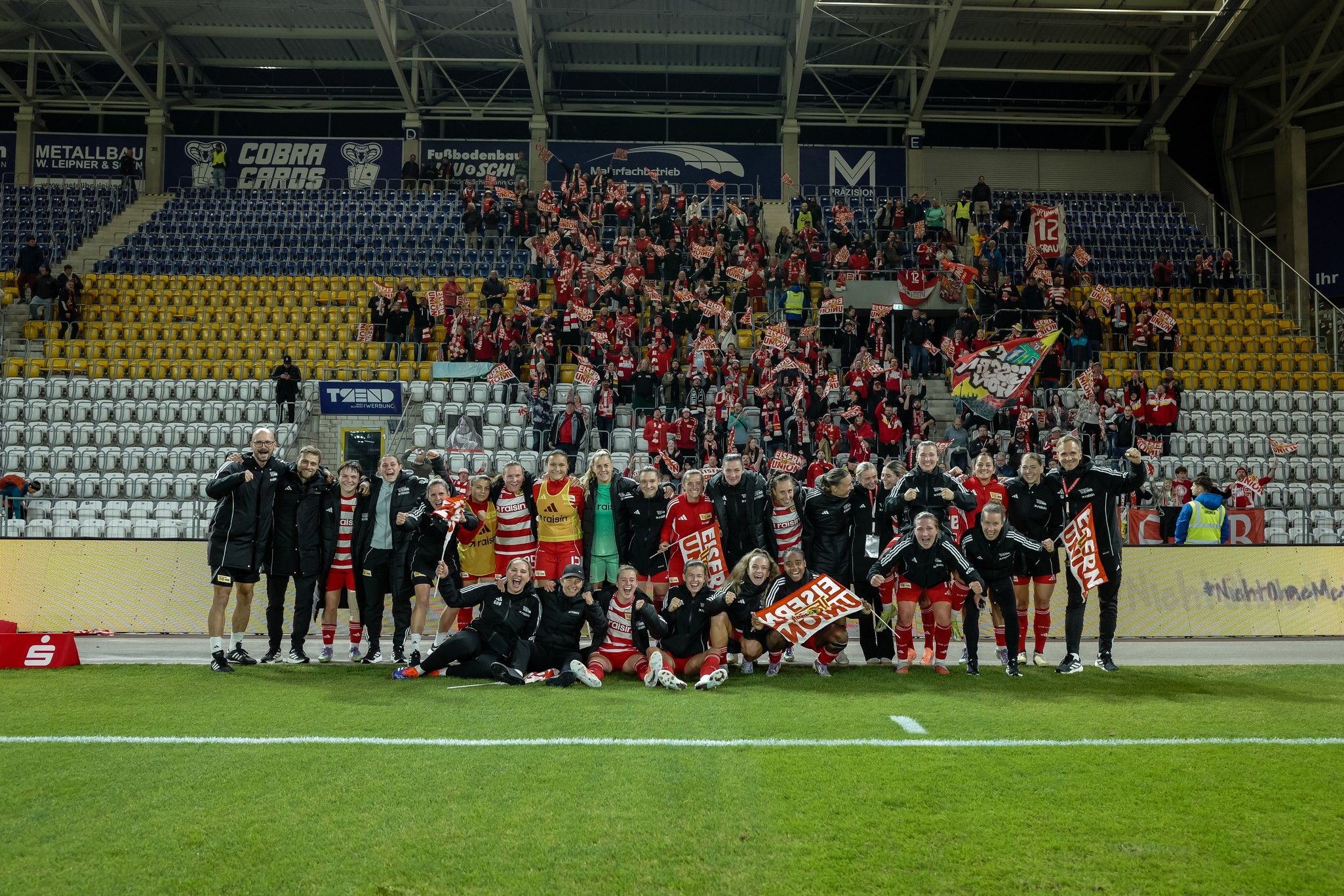 Feiernde Fußballmannschaft mit Fans im Stadion, die Banner und Schals halten. Emotionale Atmosphäre nach einem Match.