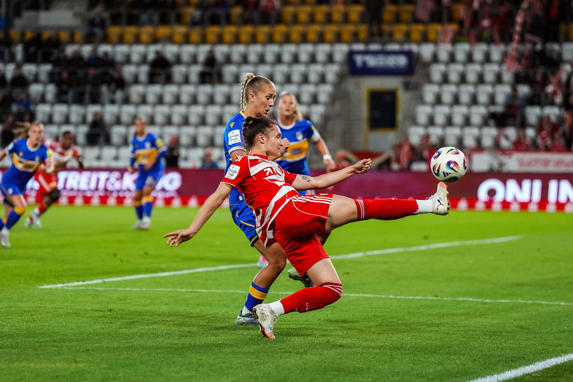 Spielszene im Fußball mit zwei Spielerinnen, eine in rotem Trikot, die den Ball mit einem hohen Beinspiel trifft, während die andere verteidigt.