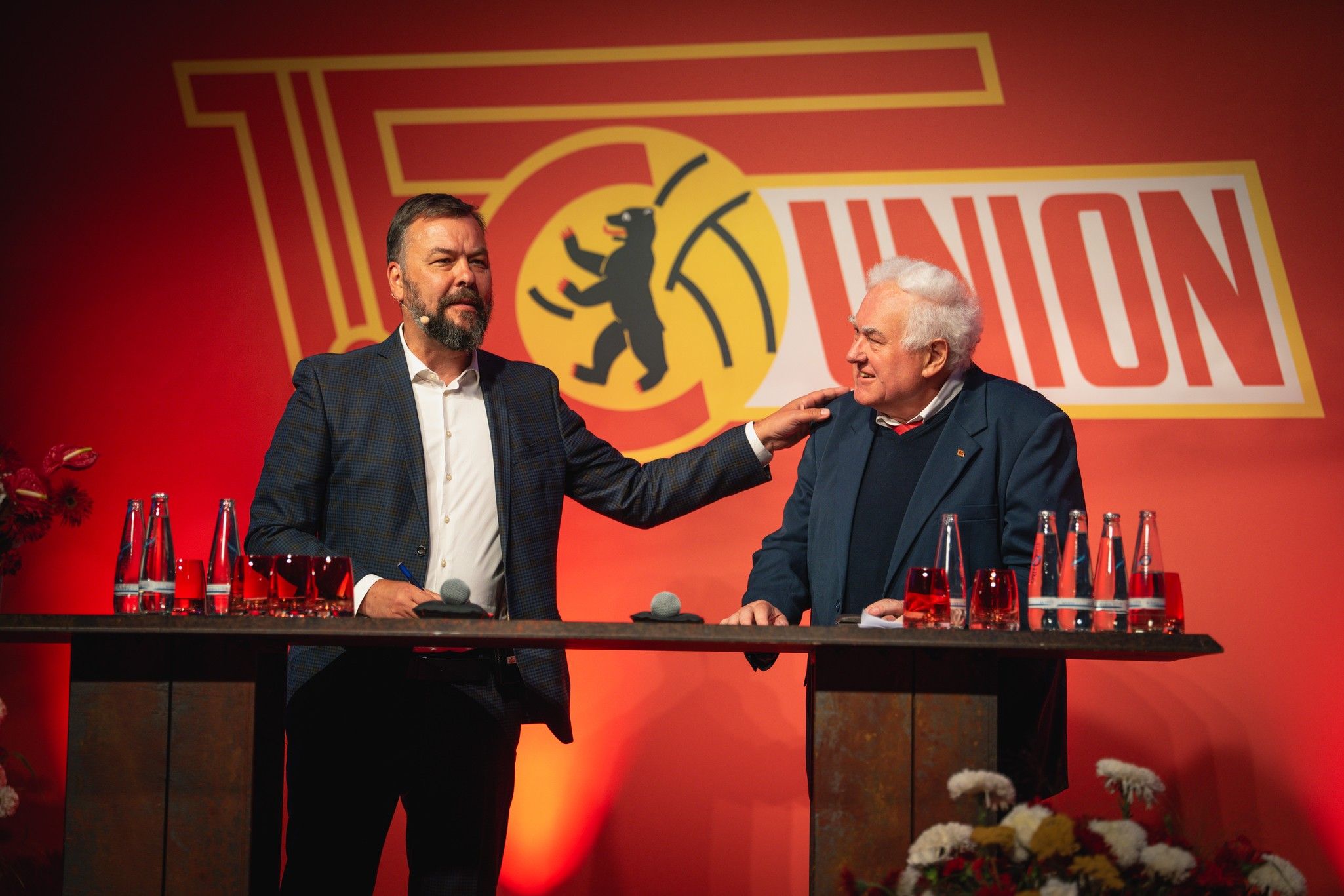 Zwei Männer stehen an einem Tisch mit Getränken, vor einer rot-gelben Hintergrundgestaltung mit dem Logo von Union Berlin.