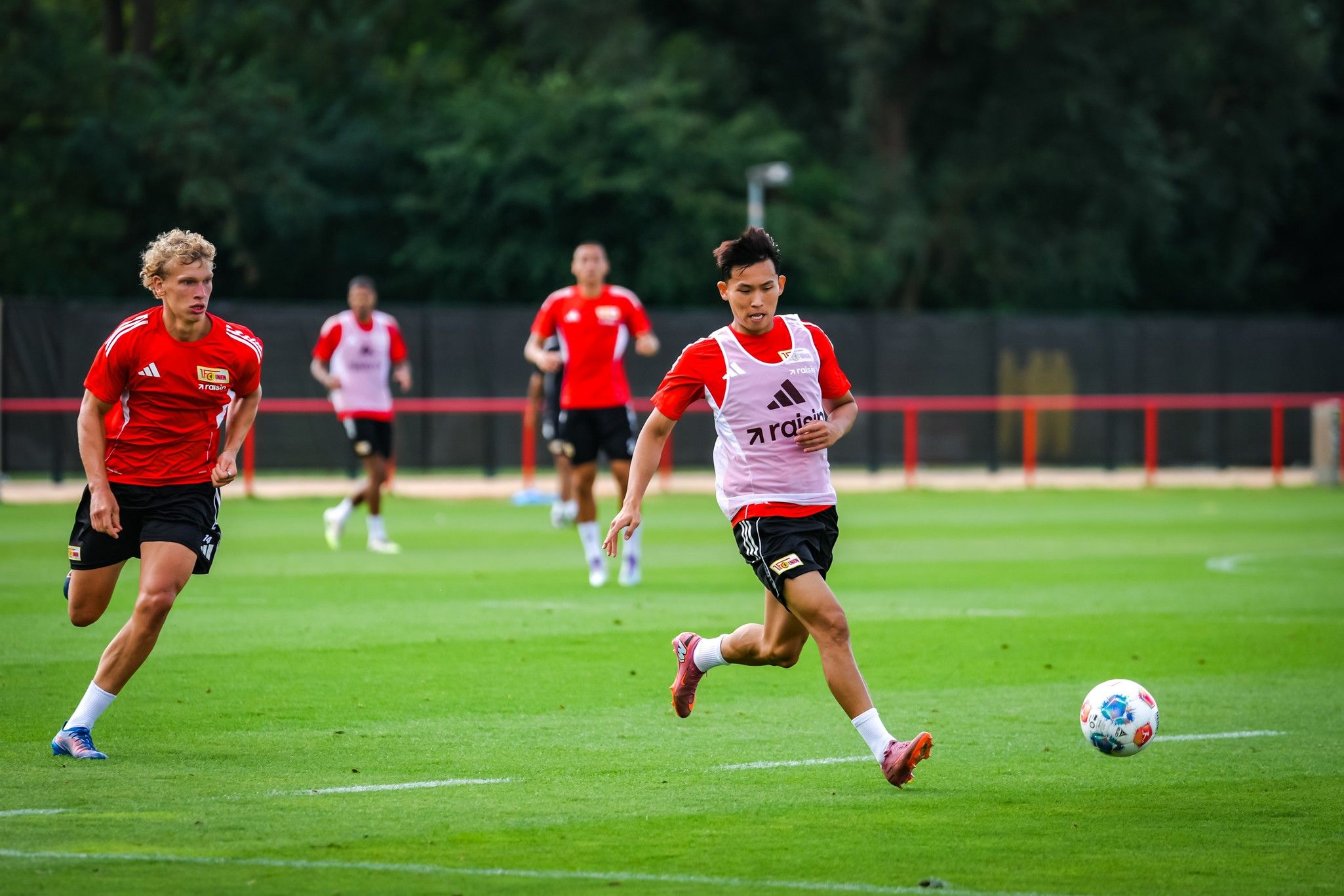 Zwei Fußballspieler in roten Trikots trainieren auf einem grünen Platz, während ein Spieler den Ball spielt.