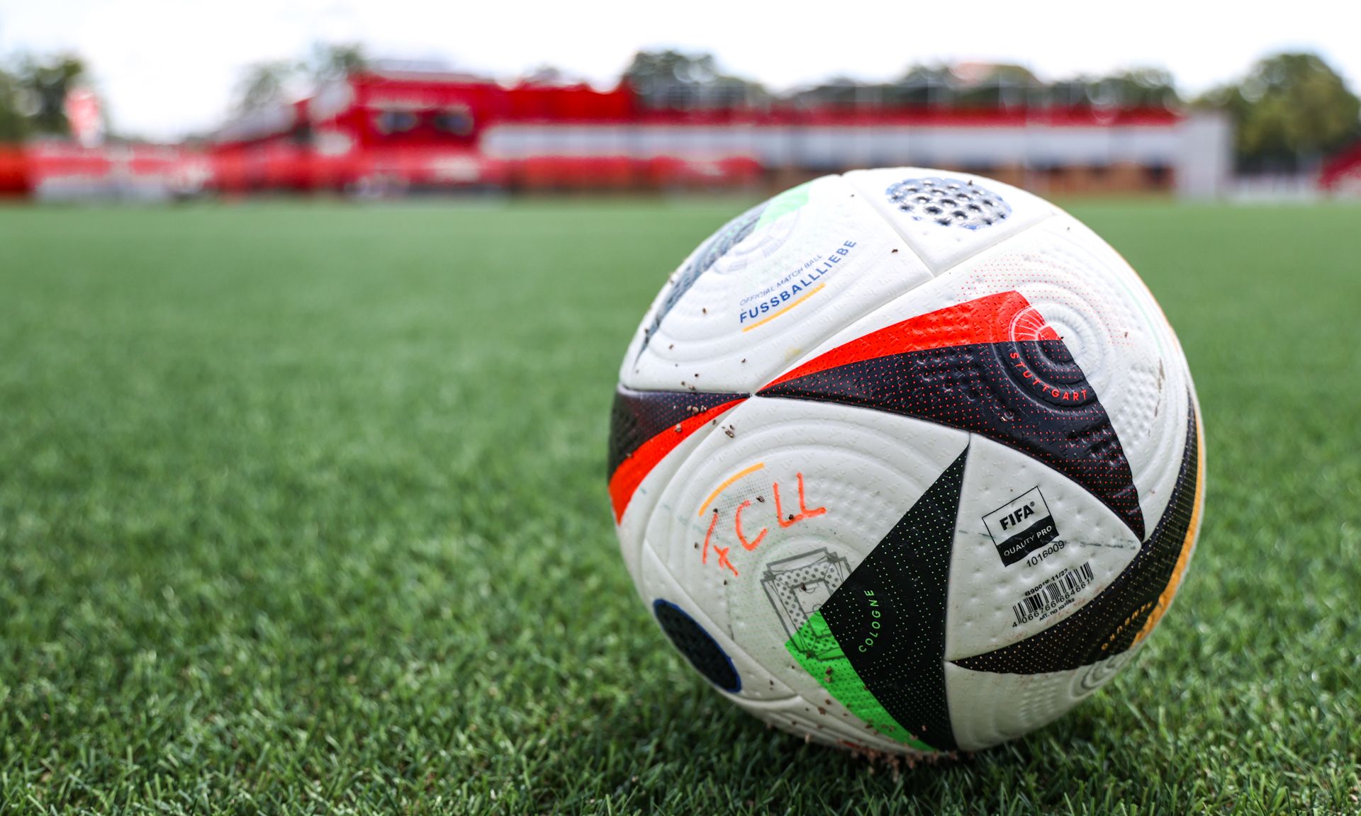 Ein Fußball mit verschiedenen Farben auf einem grünen Kunstrasenplatz, im Hintergrund verschwommen eine Sporteinrichtung.
