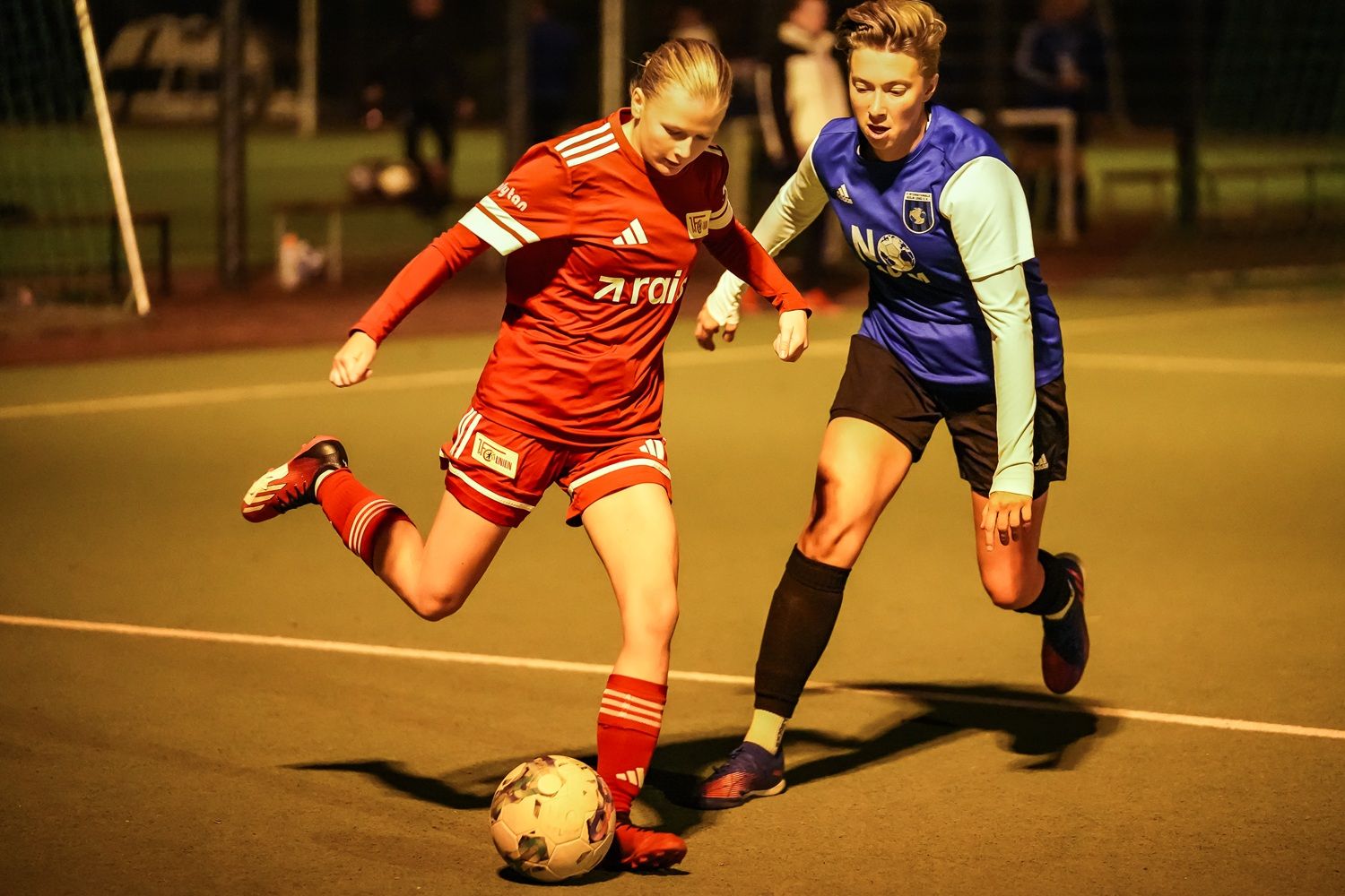 Zwei Fußballspielerinnen im Wettkampf, eine in rotem Trikot schießt den Ball, die andere in blauem Trikot versucht, sie zu verteidigen.