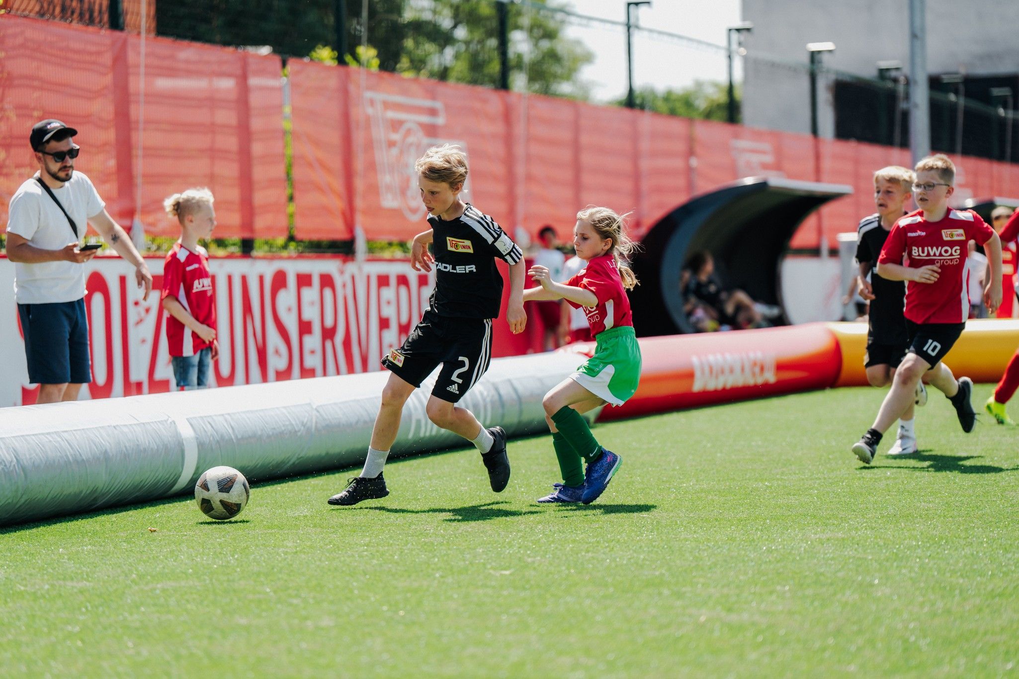 Zwei Kinder spielen Fußball auf einem Kunstrasenplatz, während weitere Kinder und ein Erwachsener zuschauen.