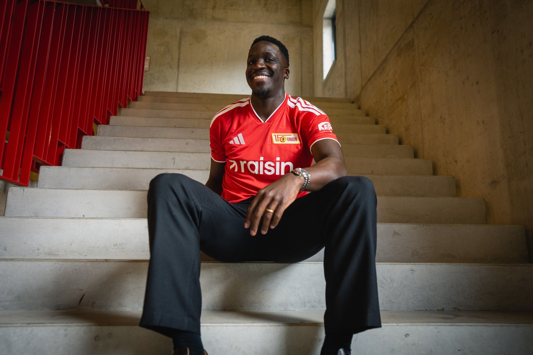 Ein lächelnder Mann in einem roten Fußballtrikot sitzt auf einer Treppe mit Betonwänden und Holzelementen im Hintergrund.
