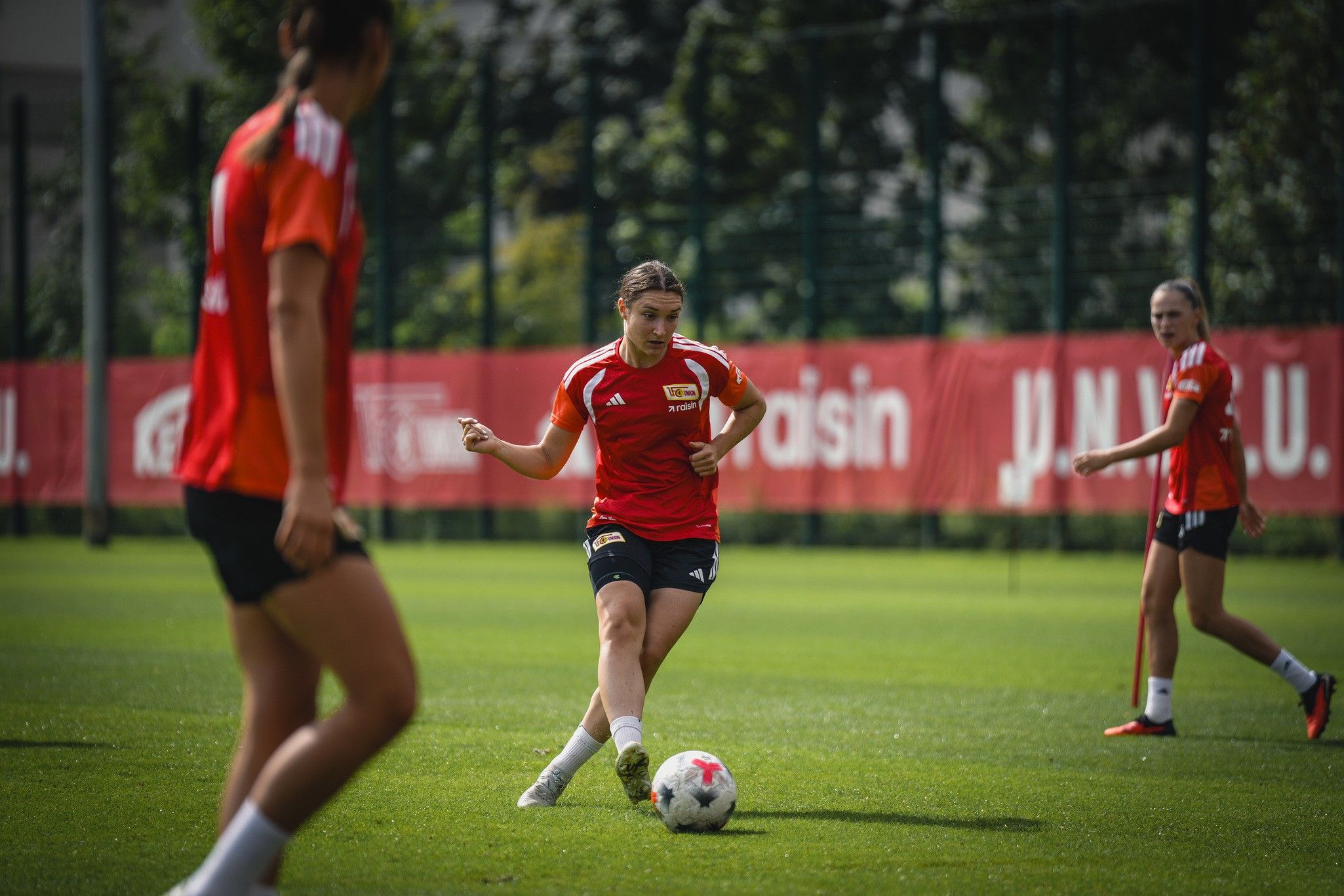 Drei Fußballspielerinnen in roten Trikots trainieren auf einem grünen Platz. Eine Spielerin führt einen Pass aus.
