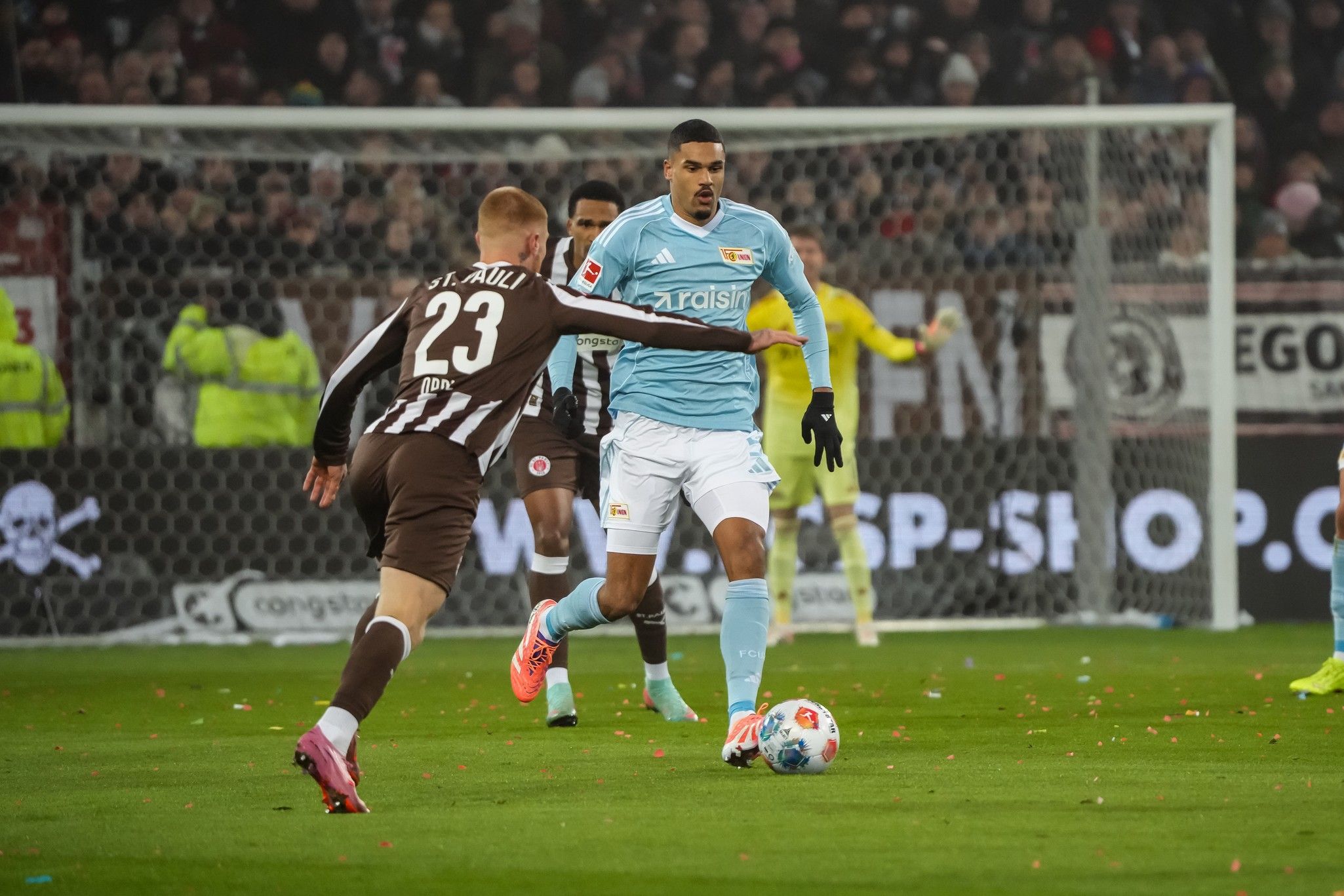 Spielszene im Fußball mit einem Spieler in hellblauem Trikot, der den Ball dribbelt, während ein Gegner ihn versucht zu stoppen.