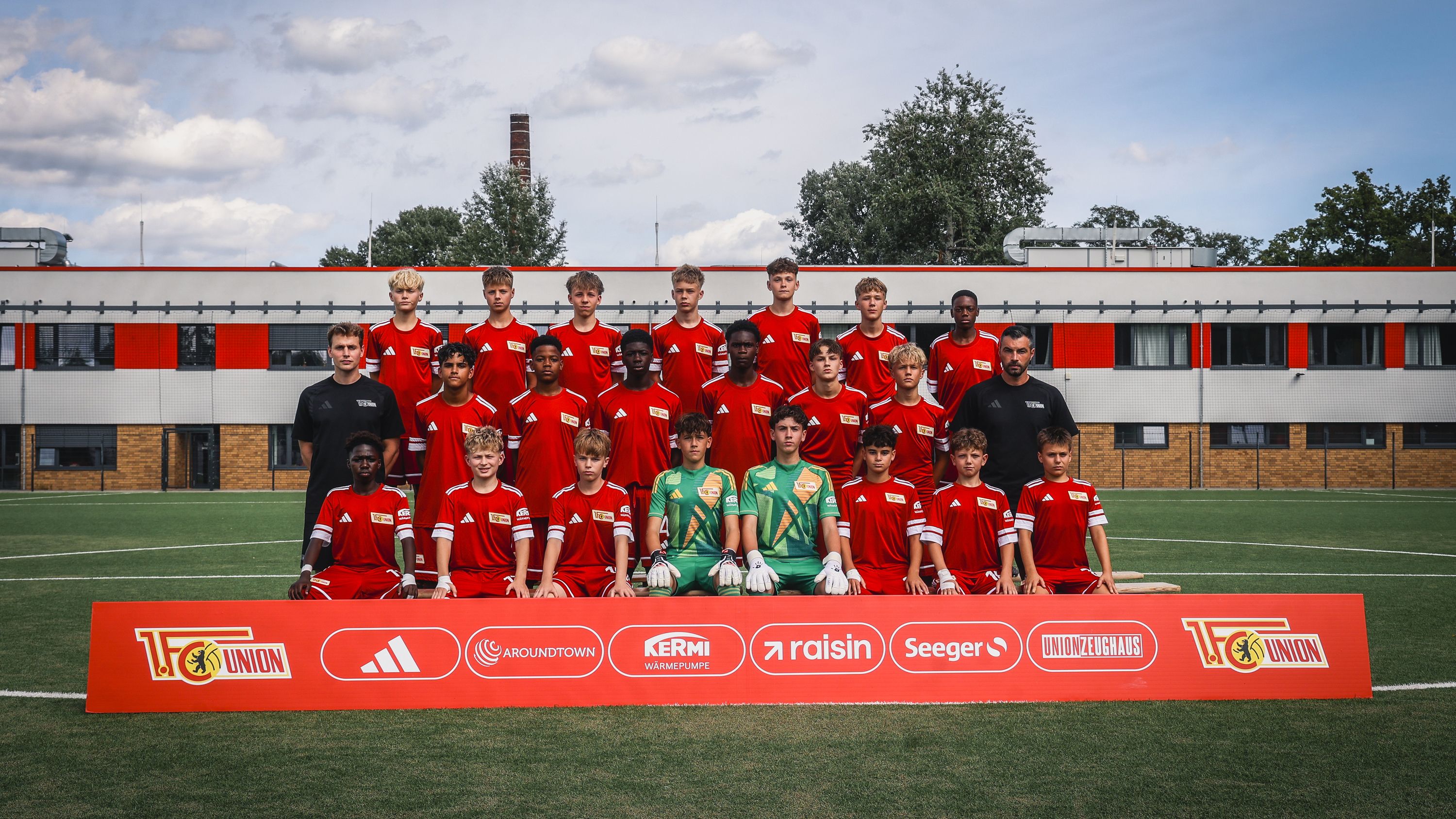 Gruppe von Jugendfußspielern in roten Trikots, posierend auf einem Fußballfeld vor einem Mannschaftsbanner.