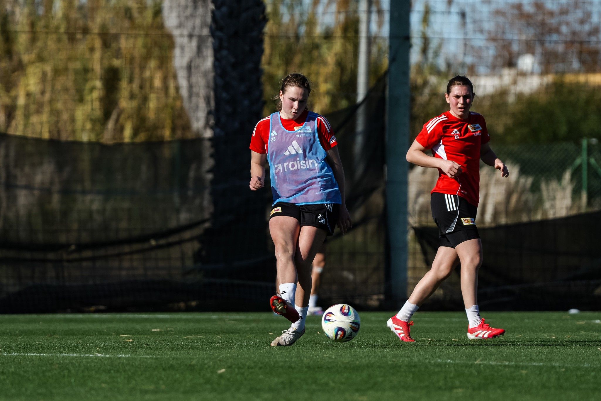 Zwei Fußballspielerinnen trainieren auf einem Rasenplatz. Eine spielt den Ball, während die andere in Bewegung ist.