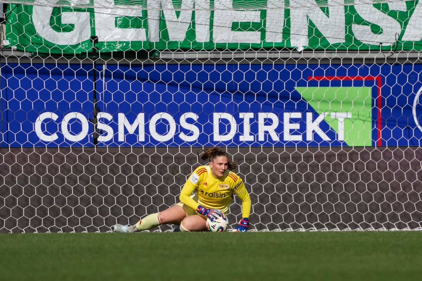 Torhüterin in gelber Ausrüstung sitzt im Tor, Blick zum Ball, Netz hinter ihr, Werbebanner im Hintergrund.