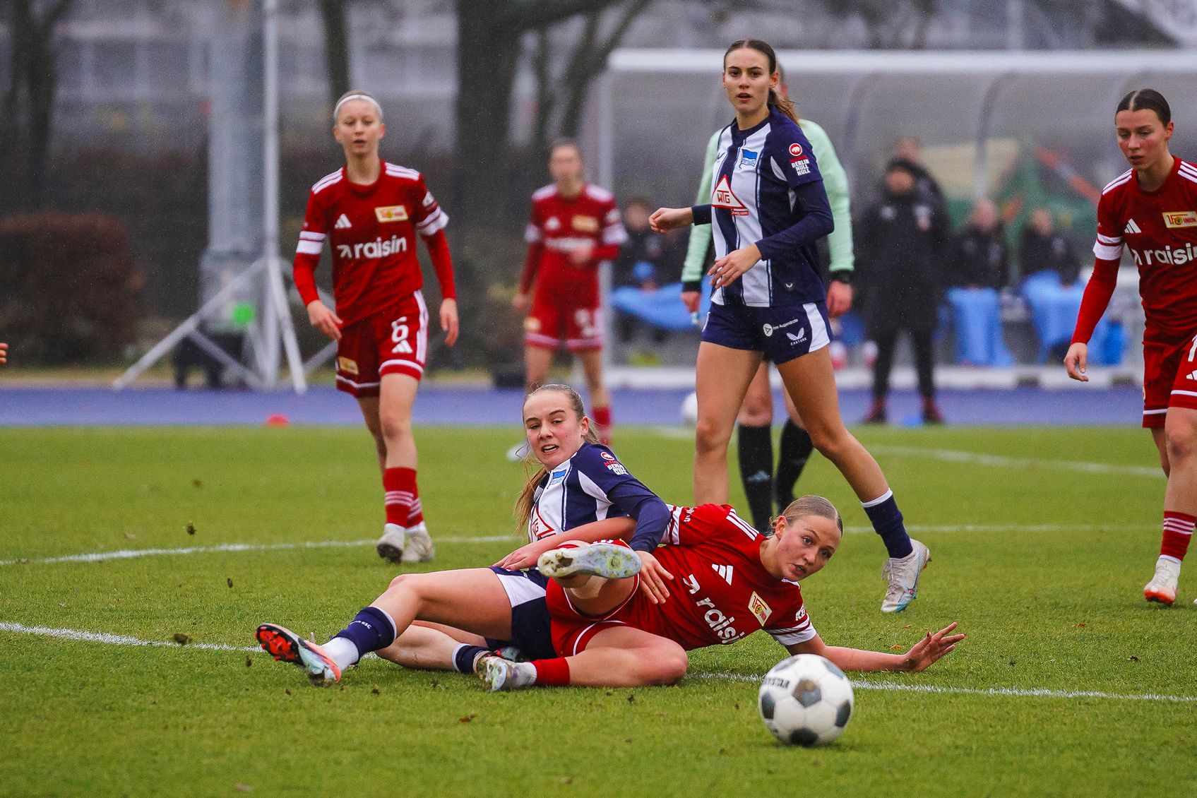 Zwei Fußballspielerinnen liegen auf dem Rasen, während andere Spielerinnen im Hintergrund stehen und das Spiel beobachten.