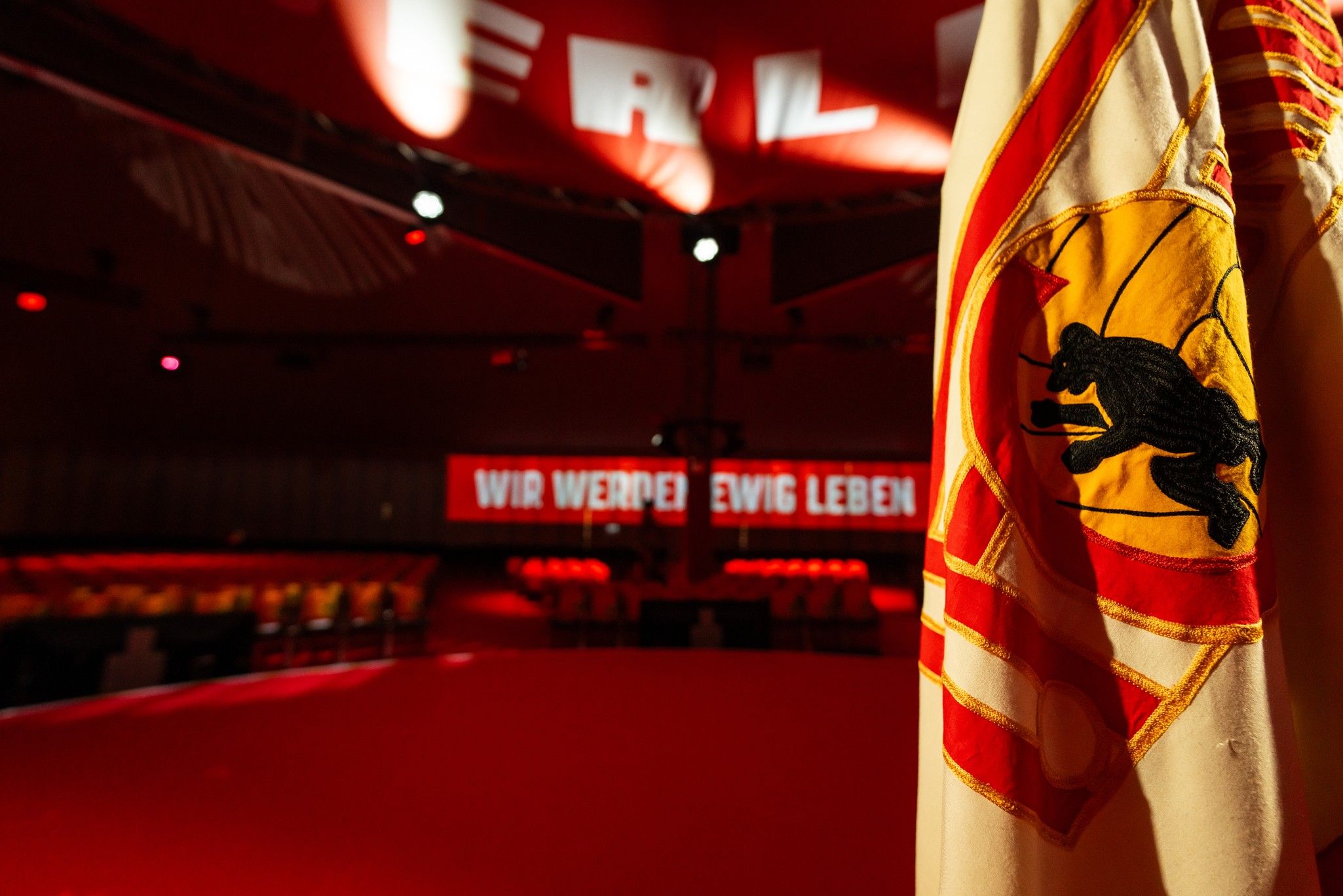 Fahne mit Wappen im Vordergrund, roter Teppich und beleuchteter Hintergrund mit dem Schriftzug "WIR WERDEN EWIG LEBEN".