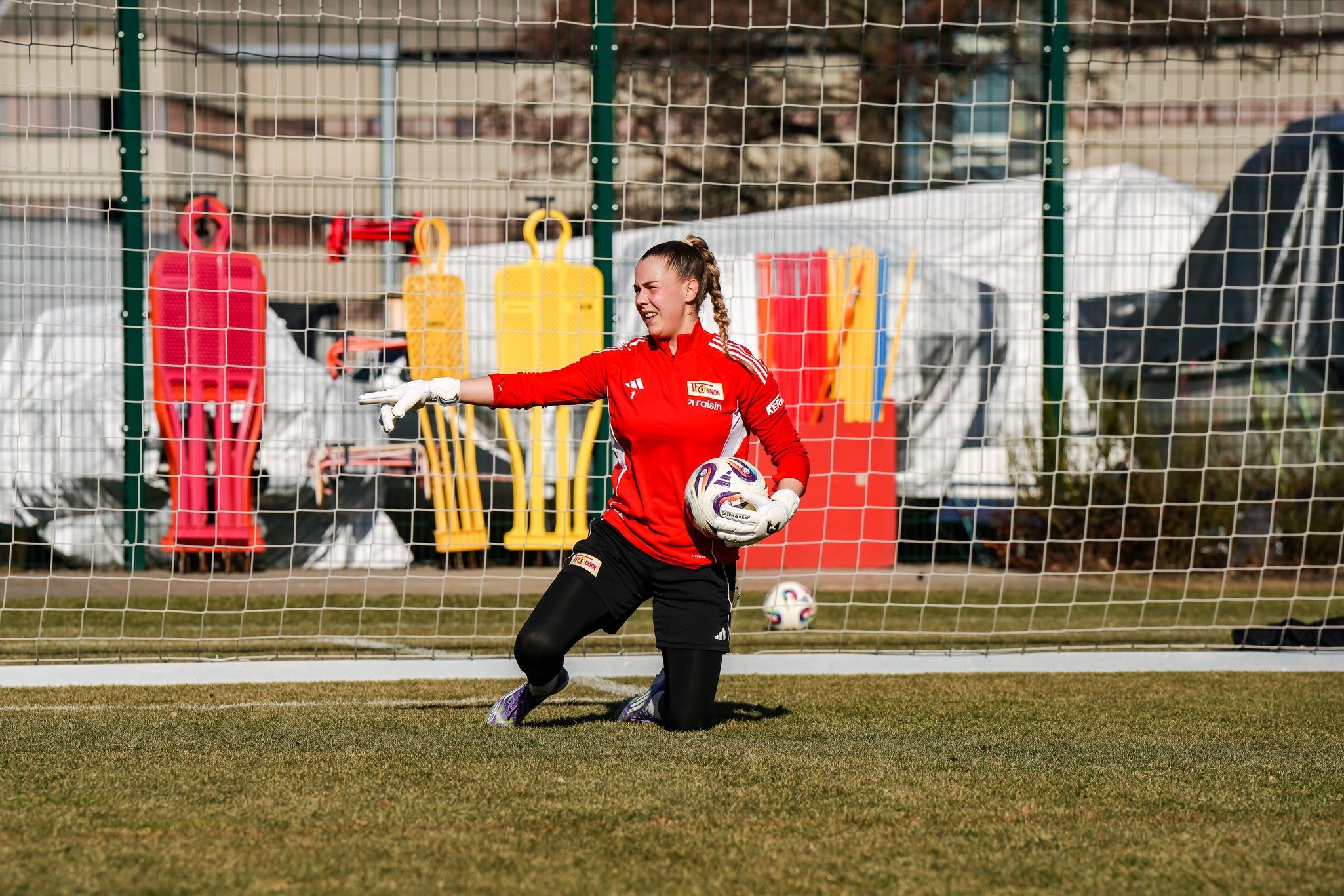 Torhüterin in rotem Trikot kniet im Torbereich und zeigt mit dem Finger auf eine Ecke, während sie einen Fußball hält.