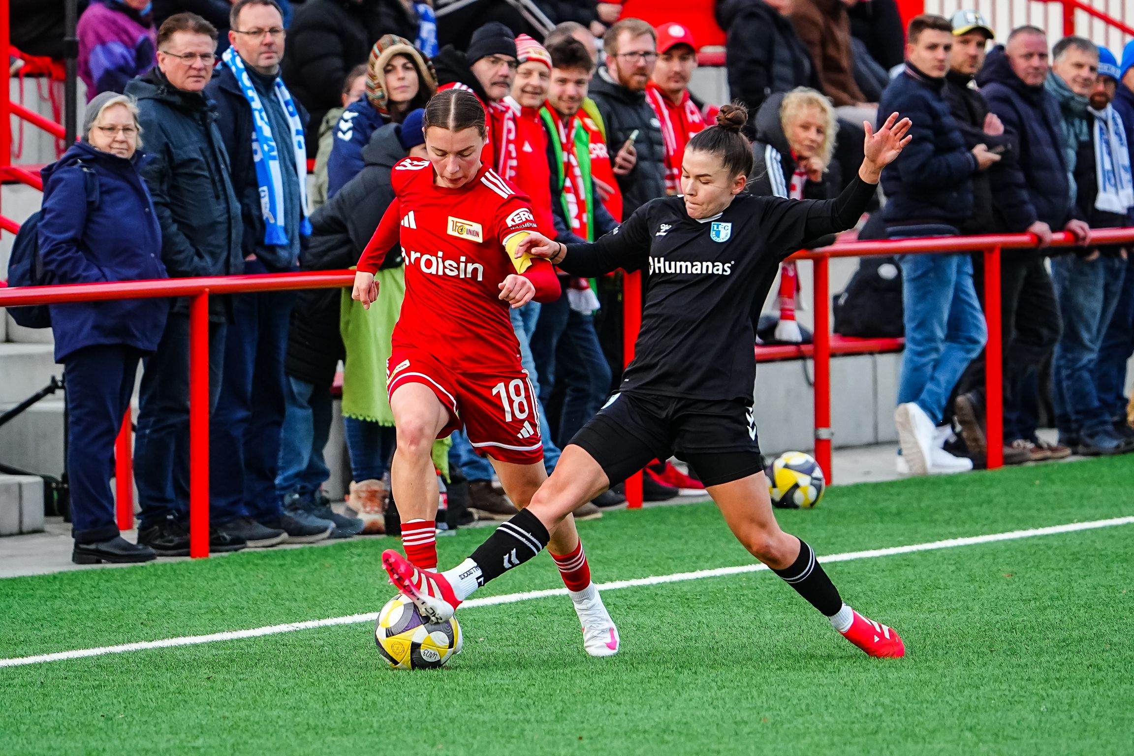 Spielszene im Frauenfußball: Eine Spielerin in roter Kleidung dribbelt den Ball, während eine Torhüterin in Schwarz den Ball defensiv anvisiert.