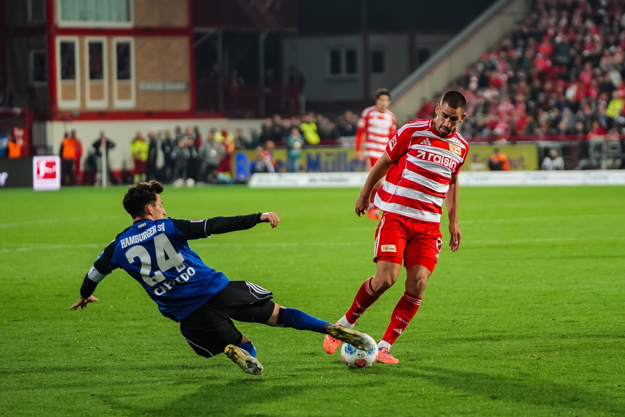 Zwei Fußballspieler im Wettkampf um den Ball auf dem Spielfeld, während ein Spieler fällt.