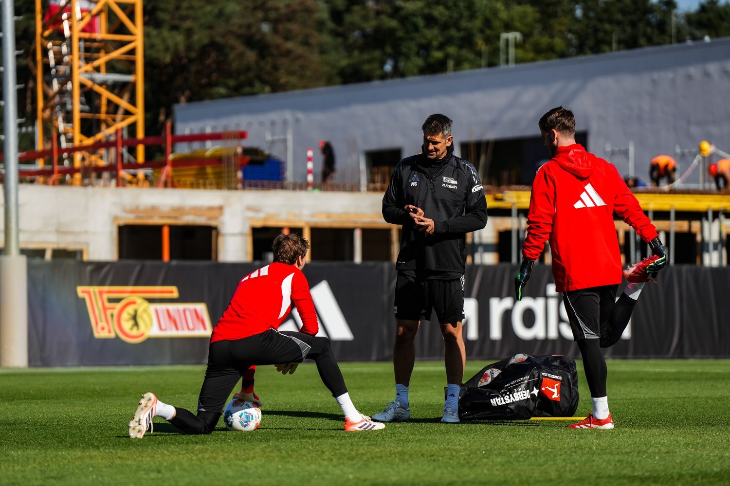 Drei Männer in Sportkleidung stehen auf einem Fußballplatz, während sie miteinander kommunizieren. Im Hintergrund sind Bauarbeiten zu sehen.