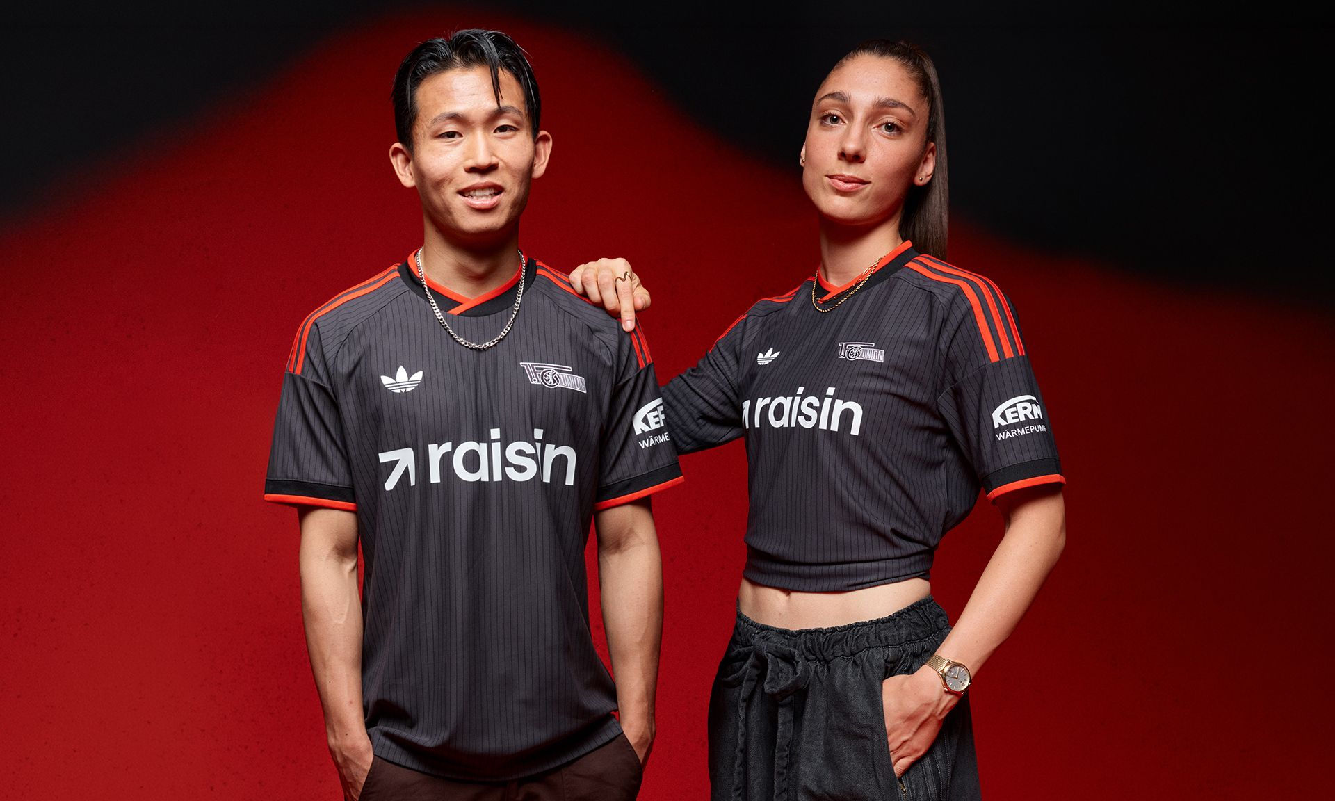 Zwei Personen in schwarzen Sporttrikots mit dem Schriftzug "raisin" vor einem roten Hintergrund.