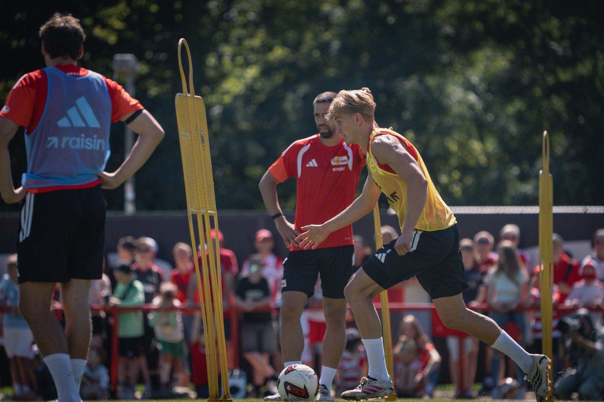 Ein Fußballspieler in gelber Weste trainiert auf dem Platz, während zwei Trainer und Zuschauer im Hintergrund zuschauen.