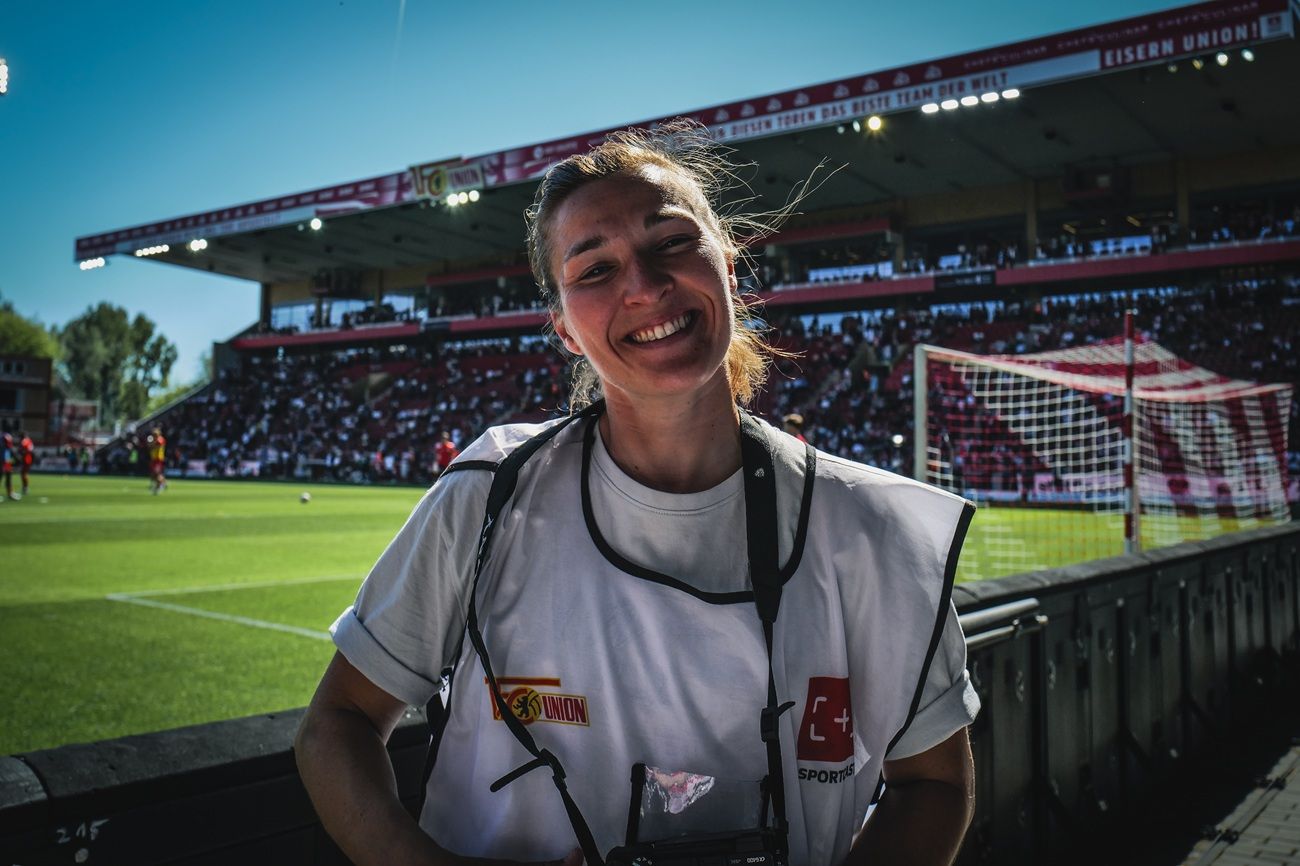 Eine lächelnde Frau in Sportbekleidung steht an einem Stadionrand, im Hintergrund ein Fußballfeld und Fans.