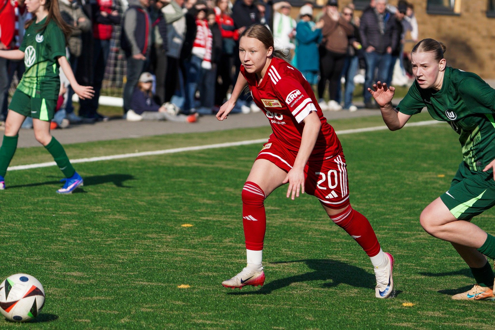Zwei Fußballspielerinnen verfolgen den Ball auf einem grünen Spielfeld, während Zuschauer im Hintergrund zuschauen.