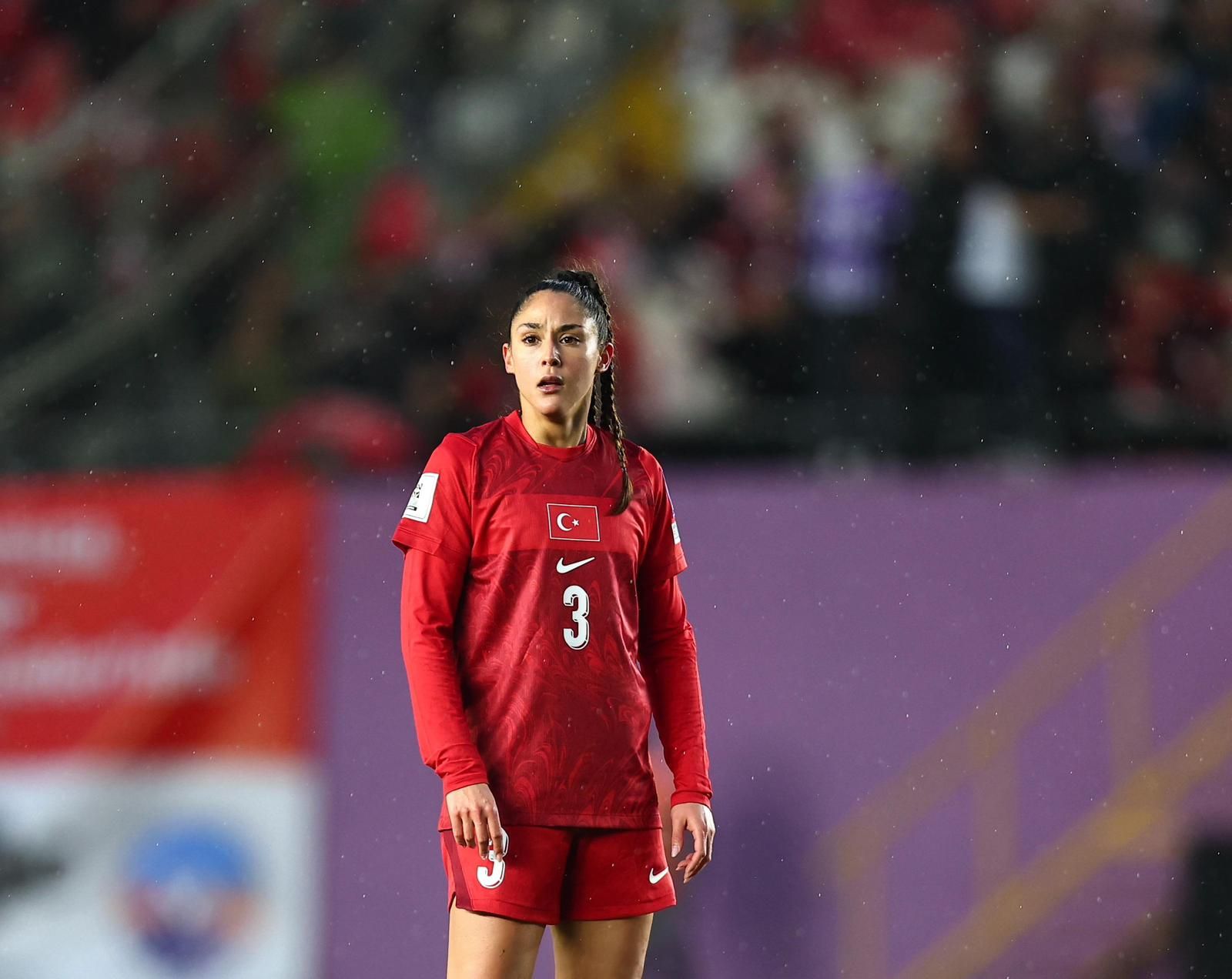 Eine Fußballspielerin in rotem Trikot mit der Nummer 3 steht auf dem Spielfeld, im Hintergrund sind verschwommene Zuschauer zu sehen.