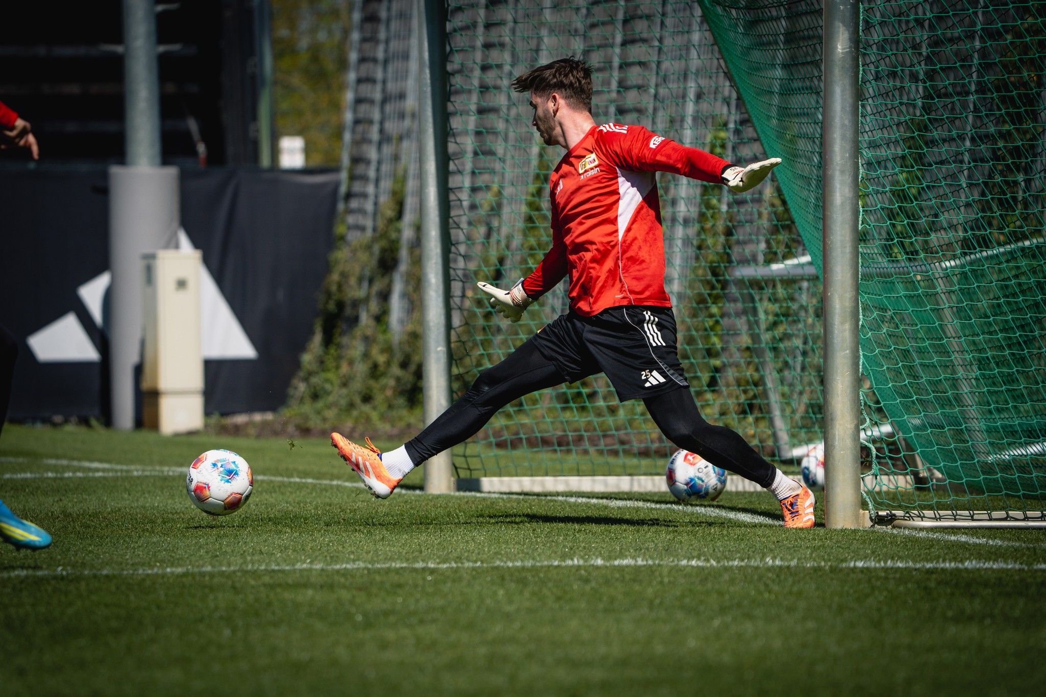 Ein Torwart in rotem Trikot springt, um einen Fußball im Training abzuwehren, während er vor einem Tor steht.