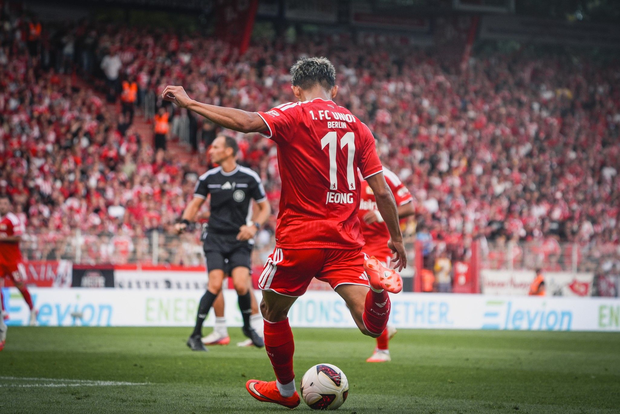 Ein Fußballspieler in rotem Trikot mit der Nummer 11 bereitet einen Schuss vor, Zuschauer im Hintergrund.