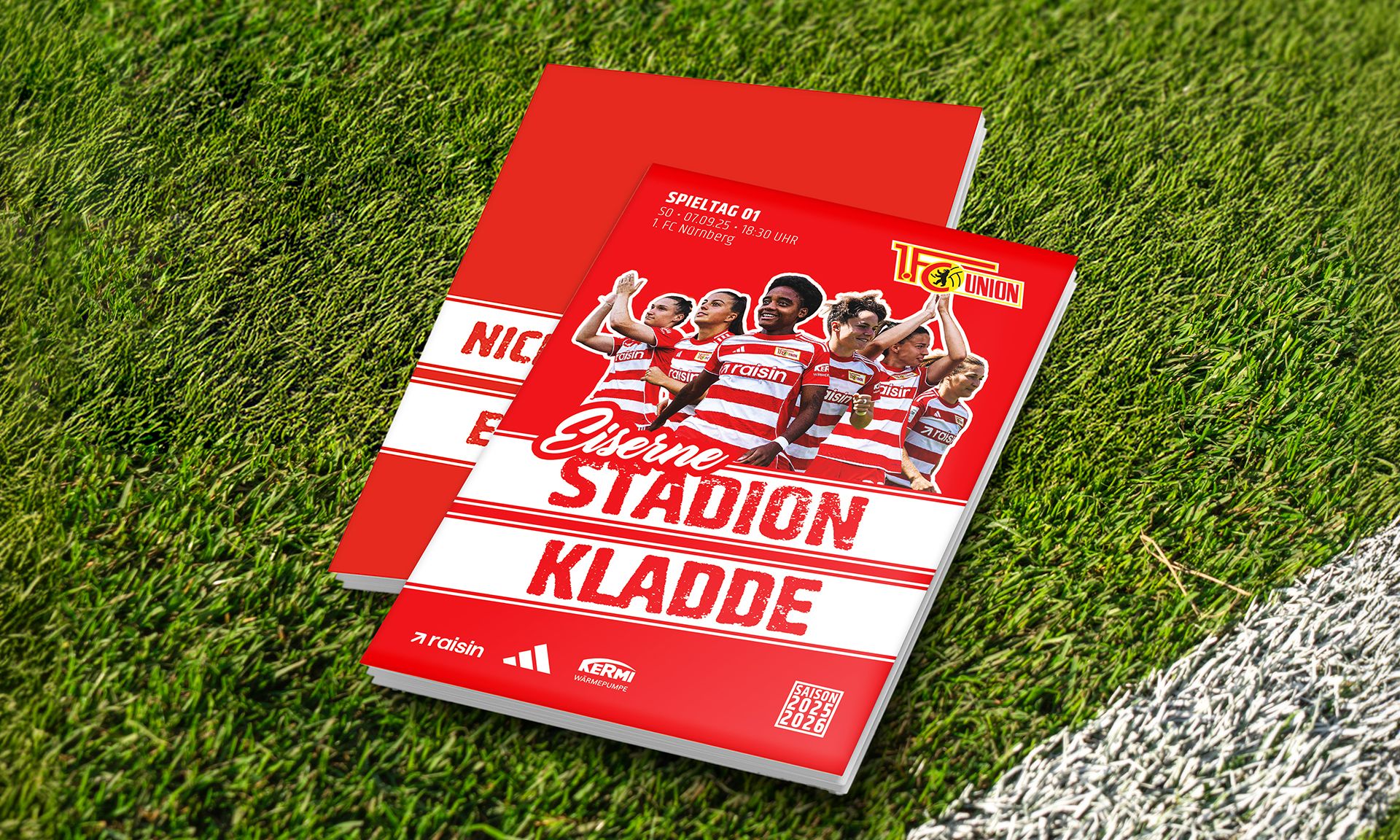 Zwei Hefte mit rotem Cover, beschriftet mit "Essener Stadion Kladde" und Fotos von Spielern, liegen auf grünem Fußballrasen.