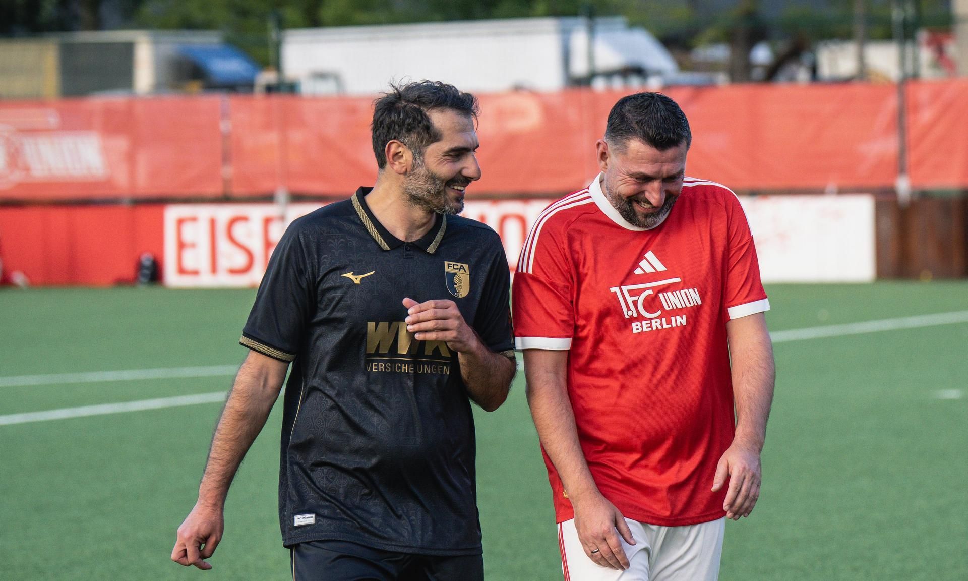 Zwei Männer in Fußballtrikots gehen lächelnd über einen Sportplatz. Hintergrund mit roten Werbeflächen.